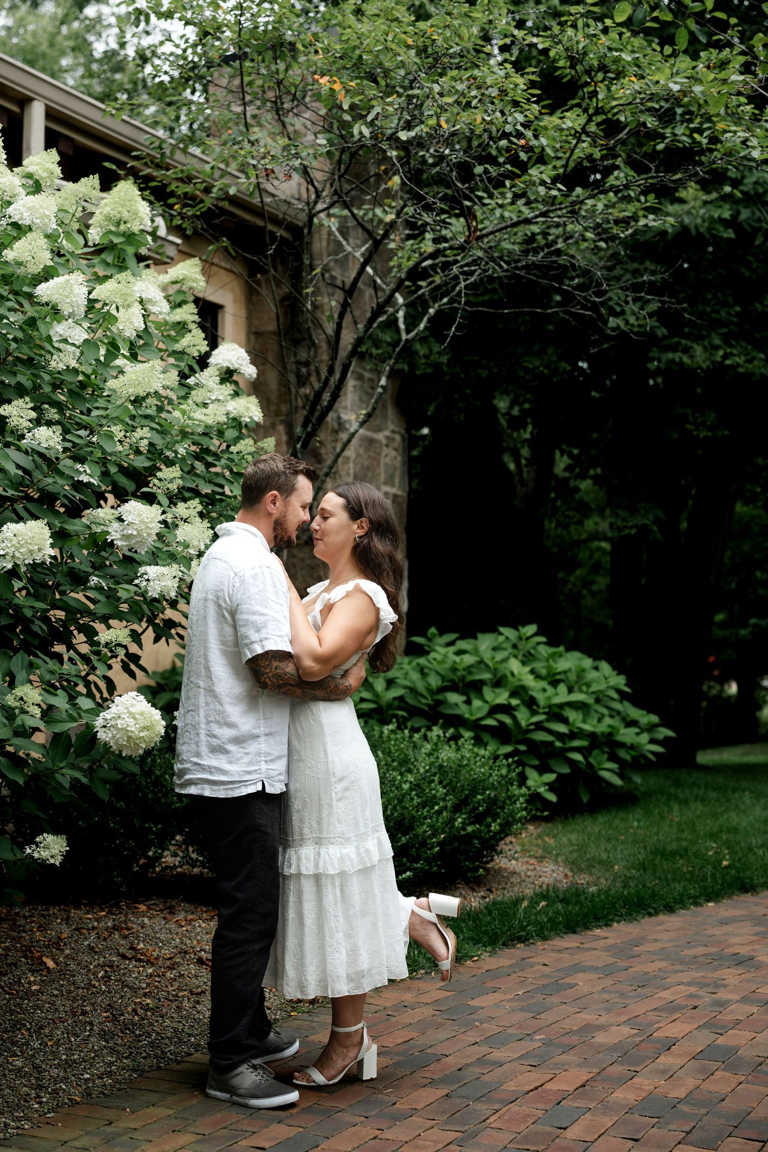Couple sharing a romantic moment in a lush garden, woman in white dress, man in white shirt, beside blooming hydrangeas.