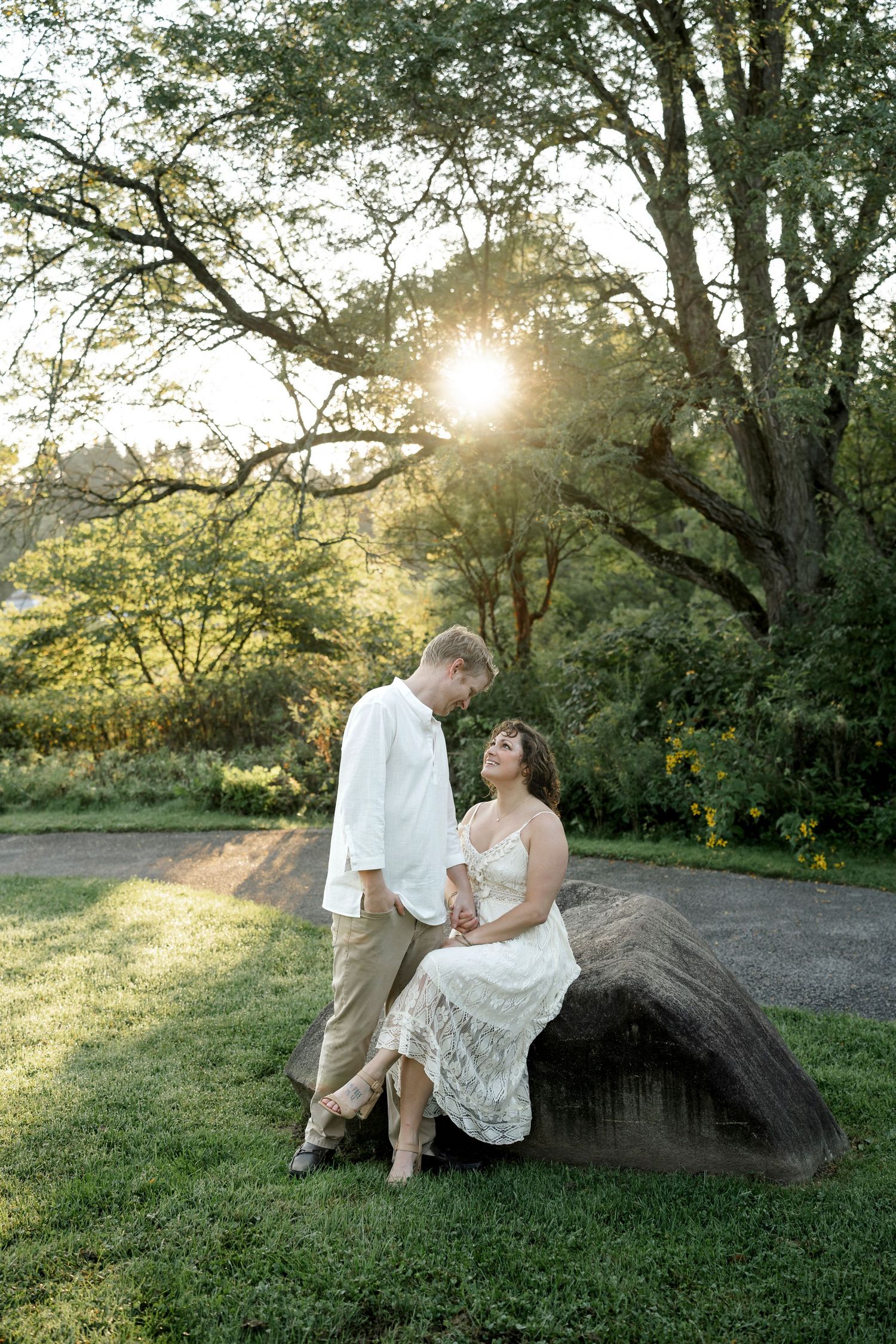 Couple in white attire sharing a romantic moment on a rock at golden hour in a lush green garden.