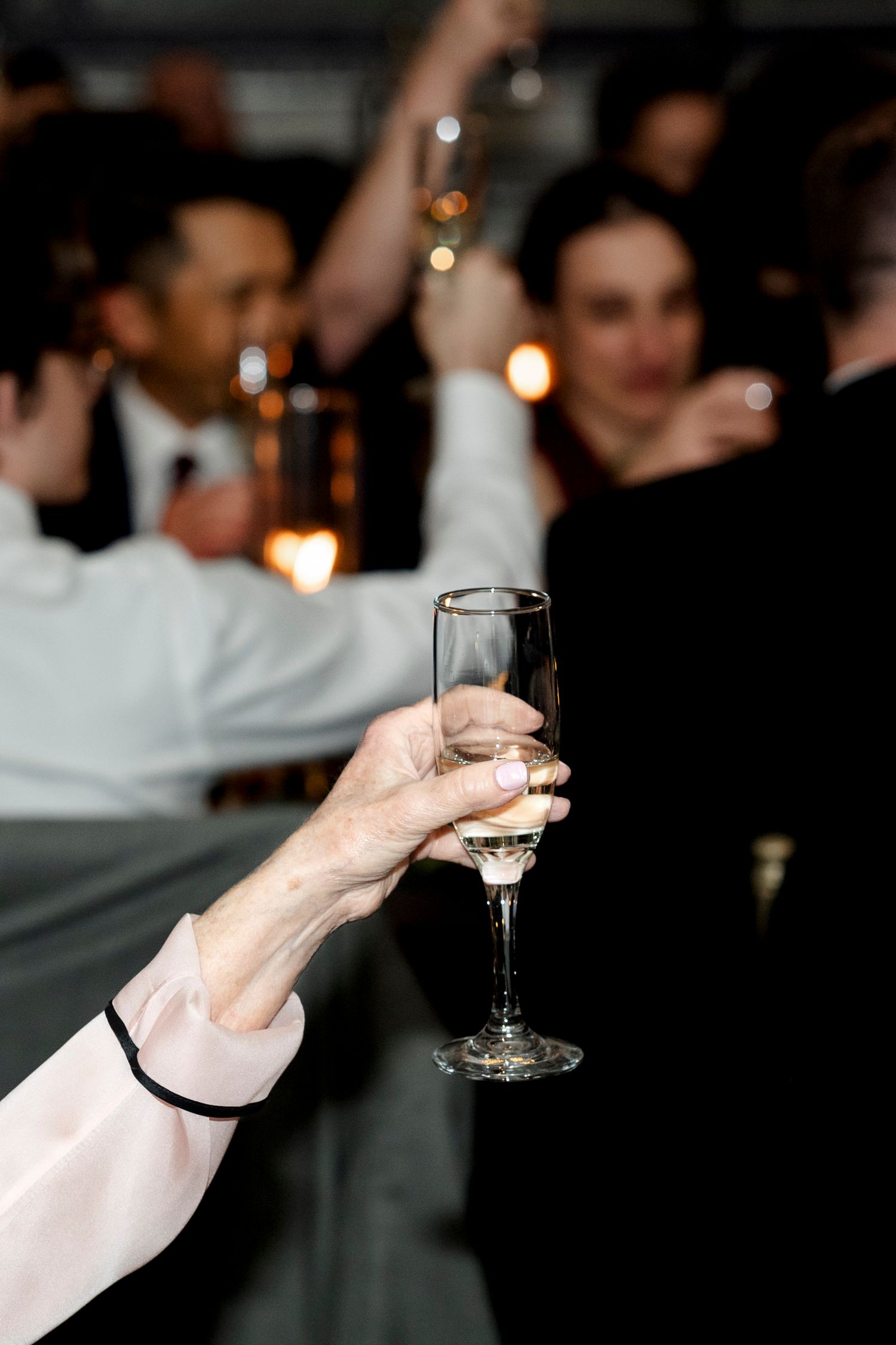 A hand in white formal attire holds up a champagne glass in a dimly lit celebration setting.