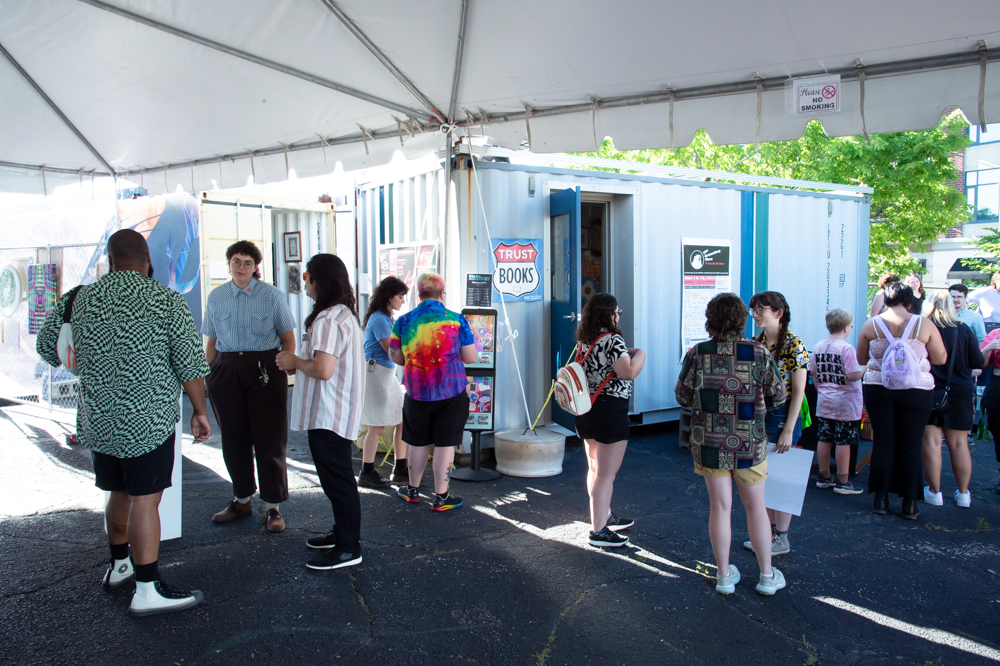 People wait in line outside white tents at an outdoor event venue during daytime.