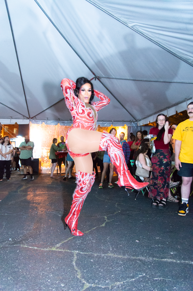 Performer in red patterned bodysuit striking dramatic pose under tent at outdoor event.