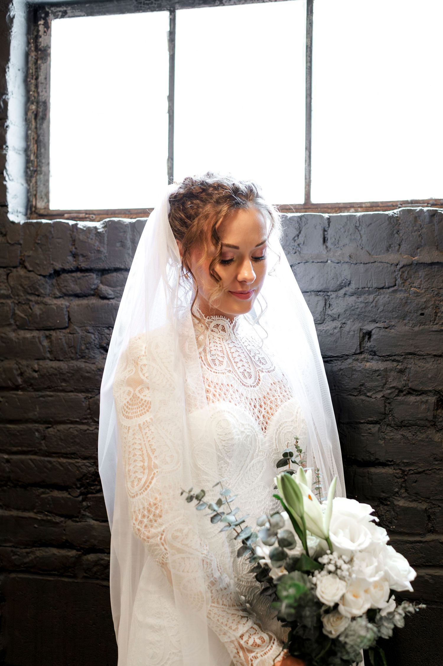 A bride wearing a lace wedding dress and veil stands in front of a window, holding a bouquet of white flowers.