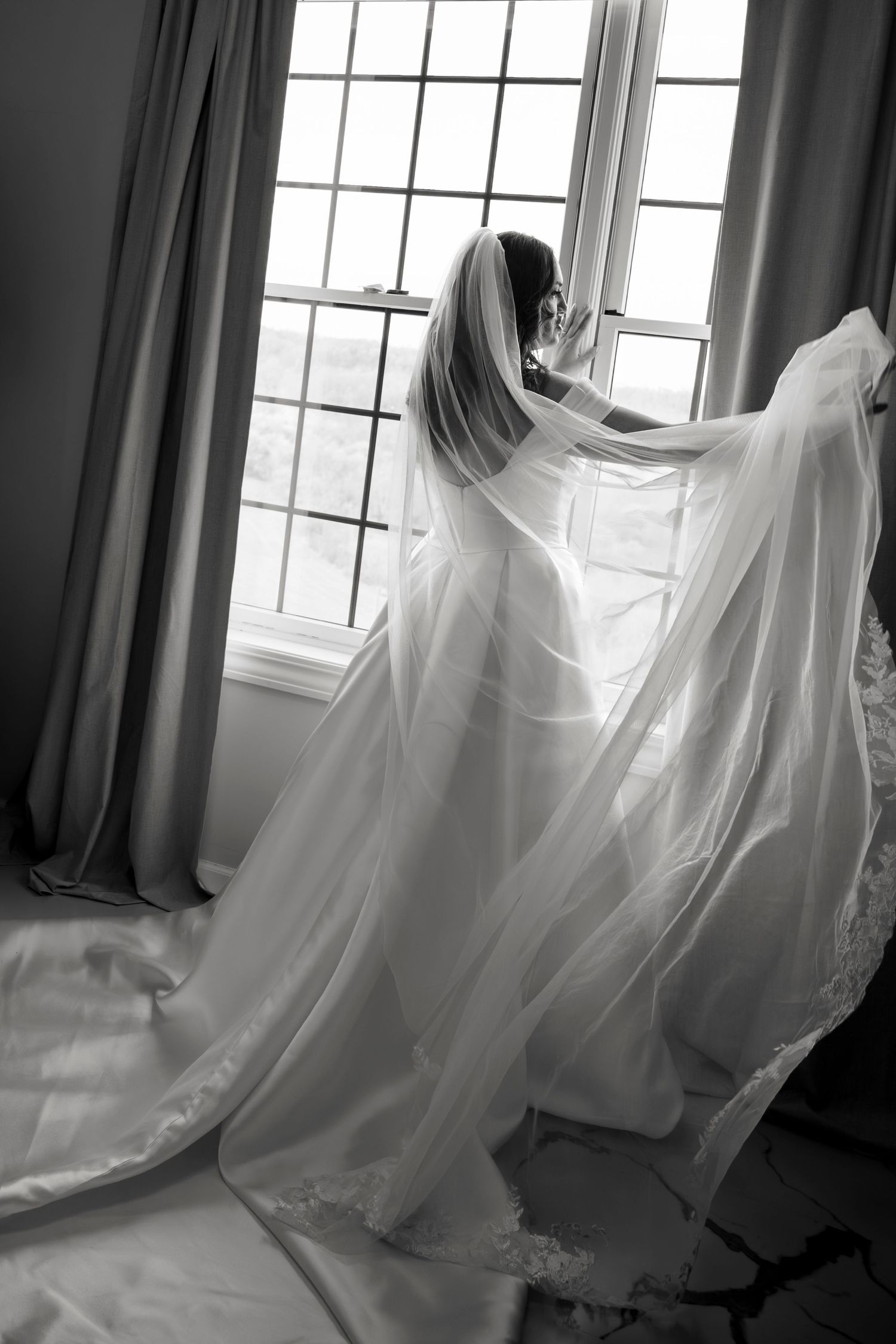 Black and white photo of a bride in a flowing wedding gown and veil standing by a large window.