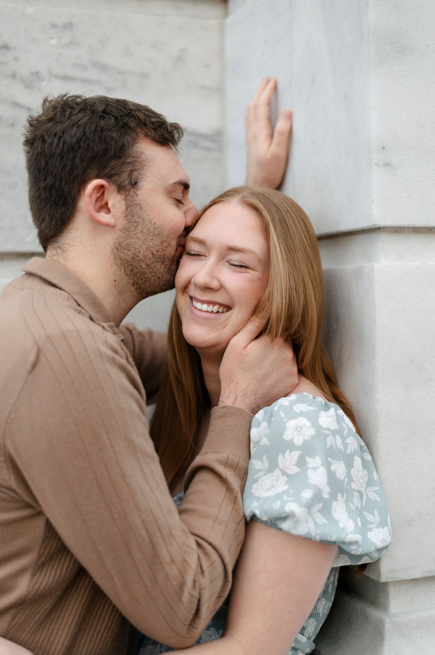 A joyful couple shares a tender moment as a man kisses a laughing woman on the cheek near a stone wall.