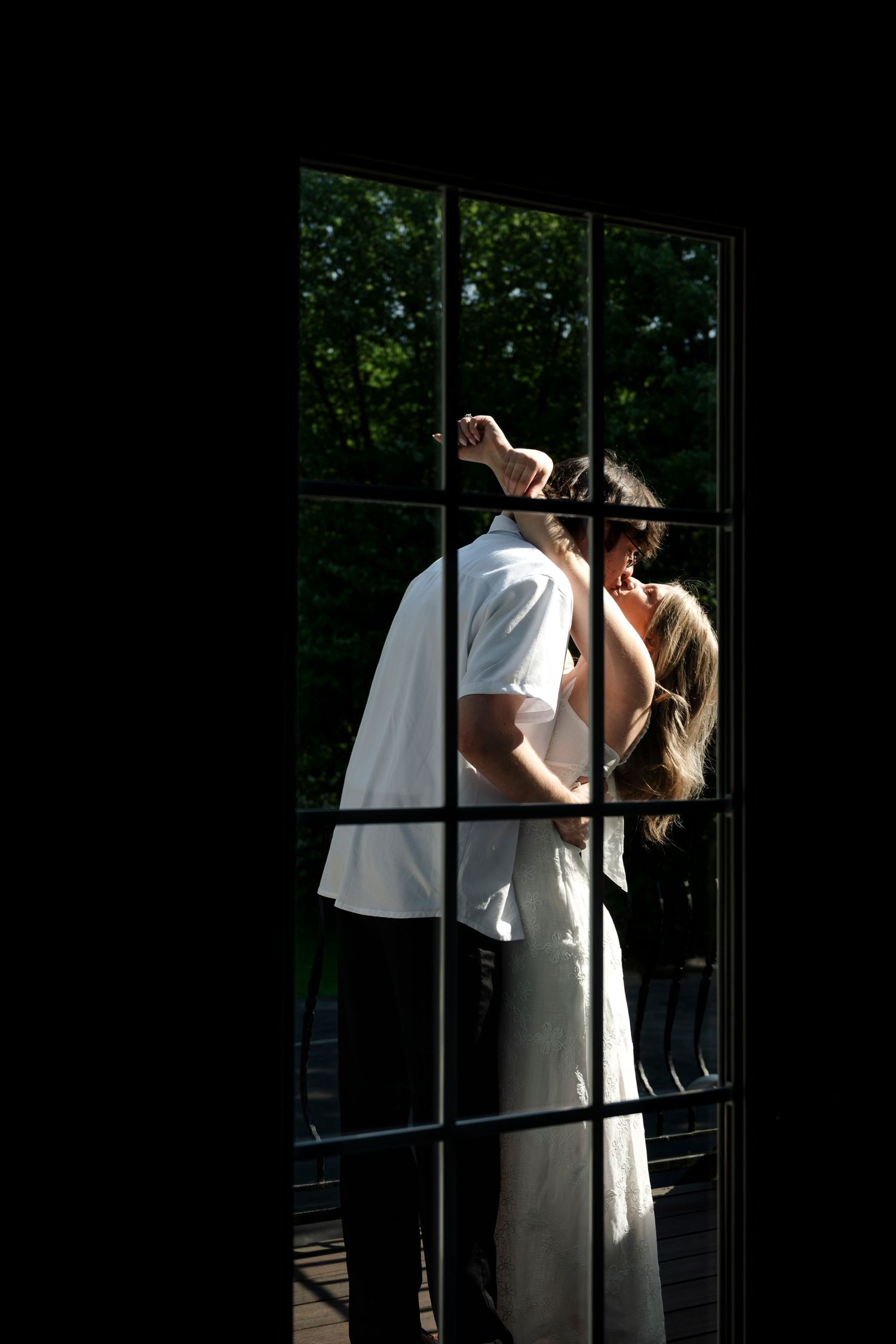 Couple sharing romantic moment dancing together viewed through glass French doors with lush green trees in background.