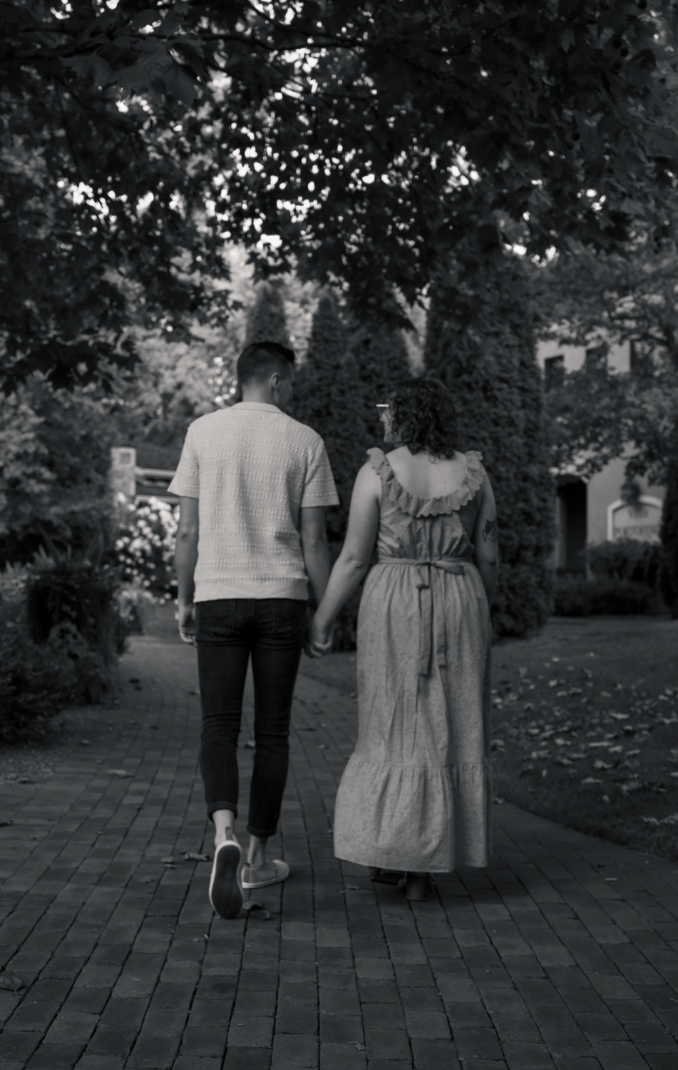 Black and white photo of a couple holding hands walking along a brick path through a lush tree-lined garden.