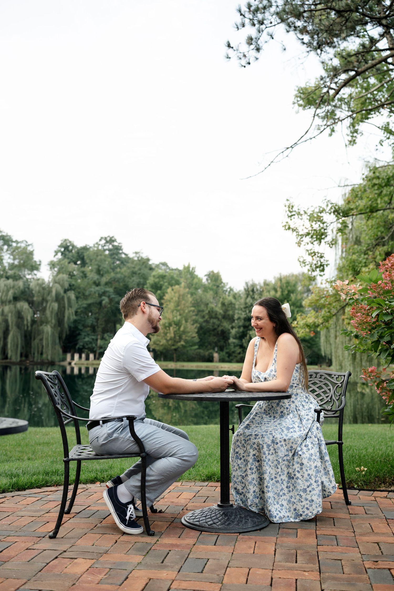 Couple holding hands at outdoor patio table by a pond, smiling at each other in a lush garden setting.