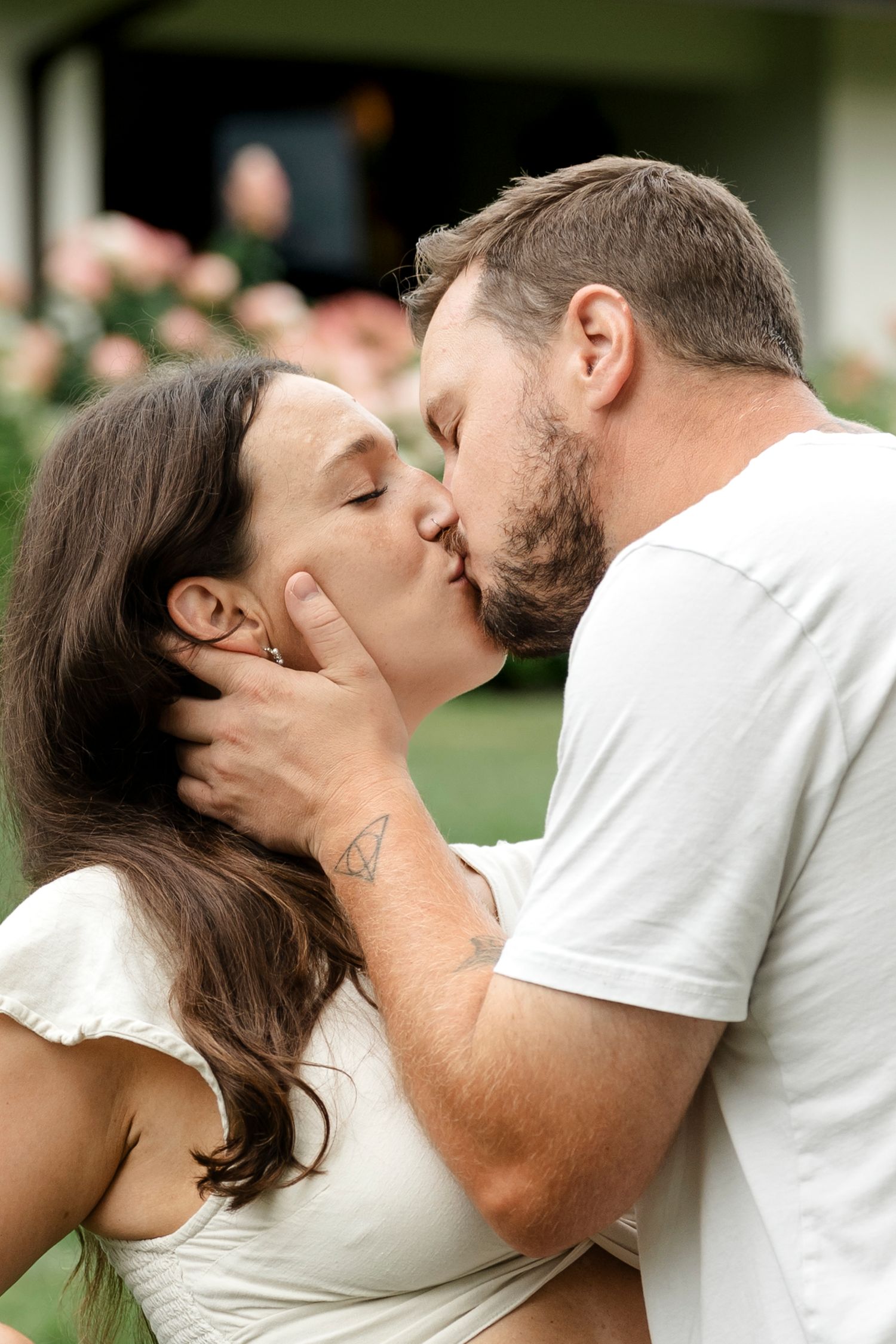 A couple shares a romantic kiss outdoors, with pink roses and greenery in the background.