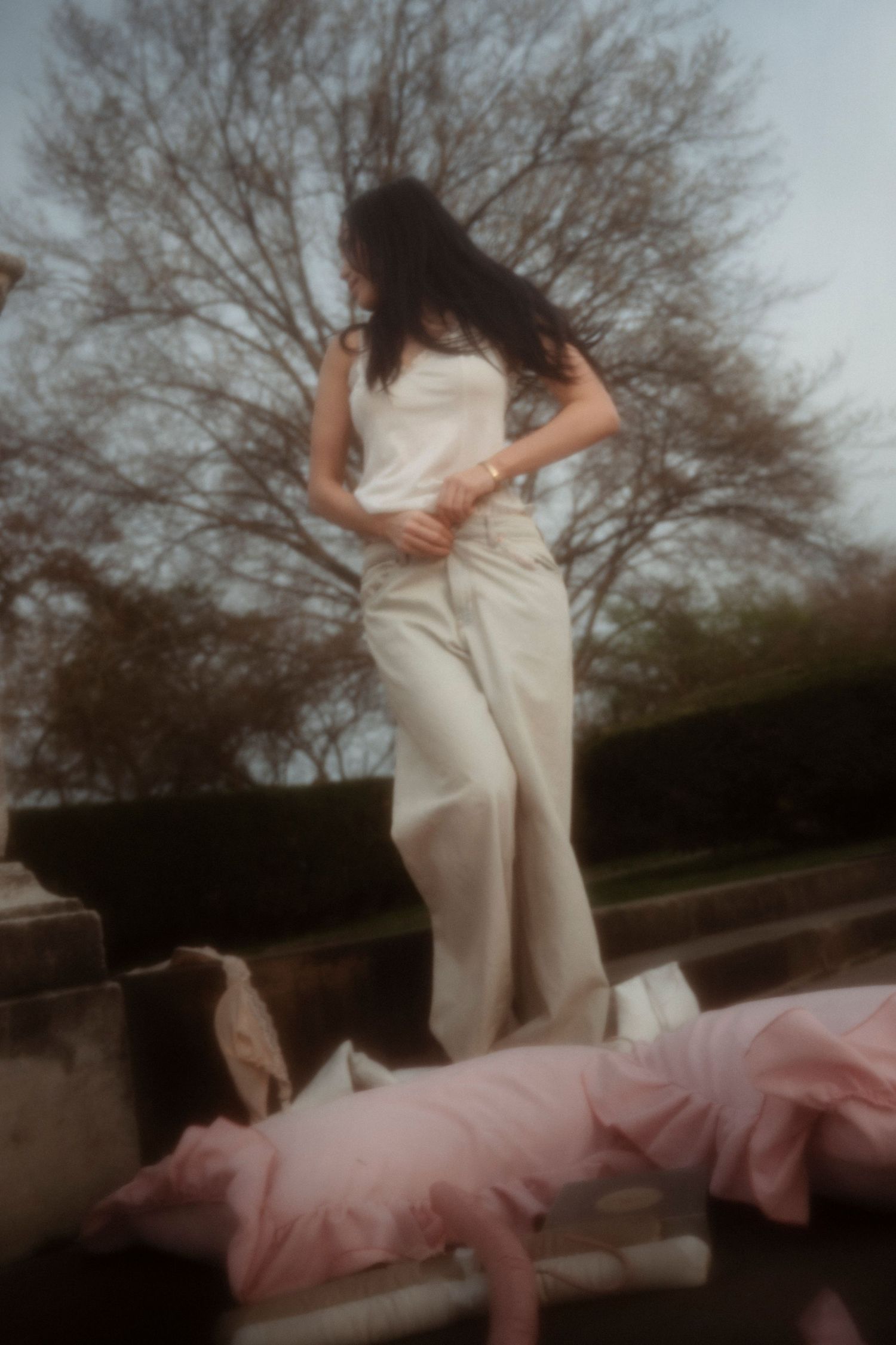 Woman in white linen outfit standing outdoors near pink pillows with bare trees in background.