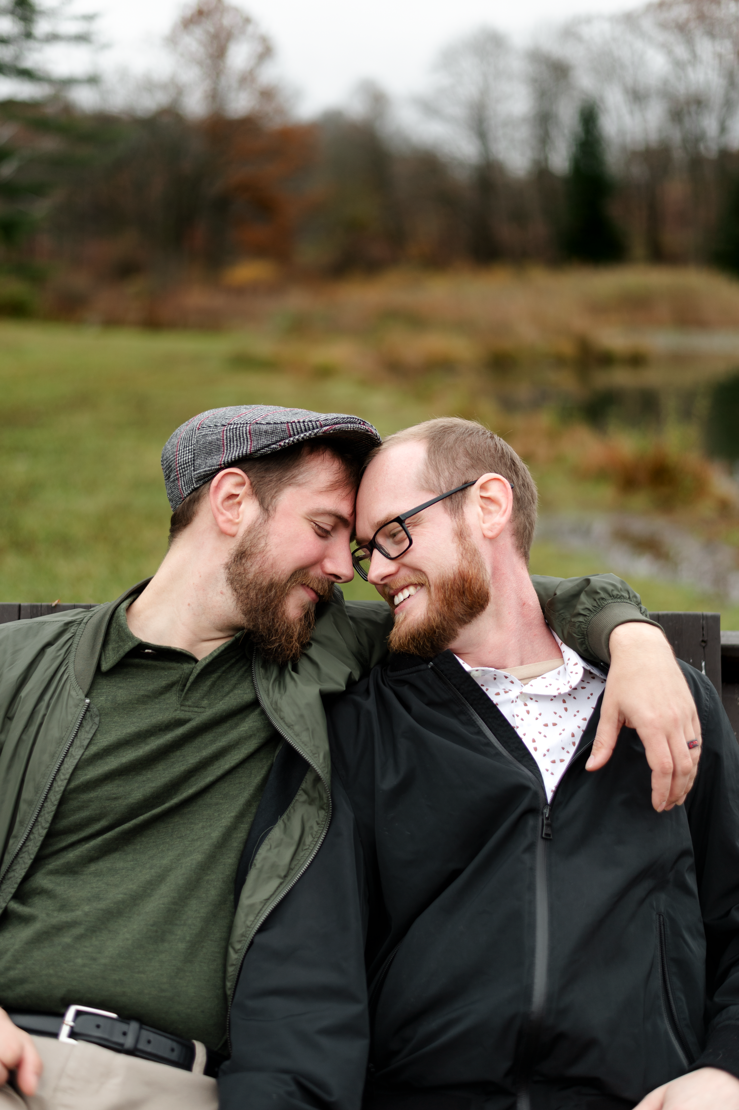 Two bearded men sharing a tender moment outdoors, touching foreheads and smiling on a bench with a green landscape behind them.