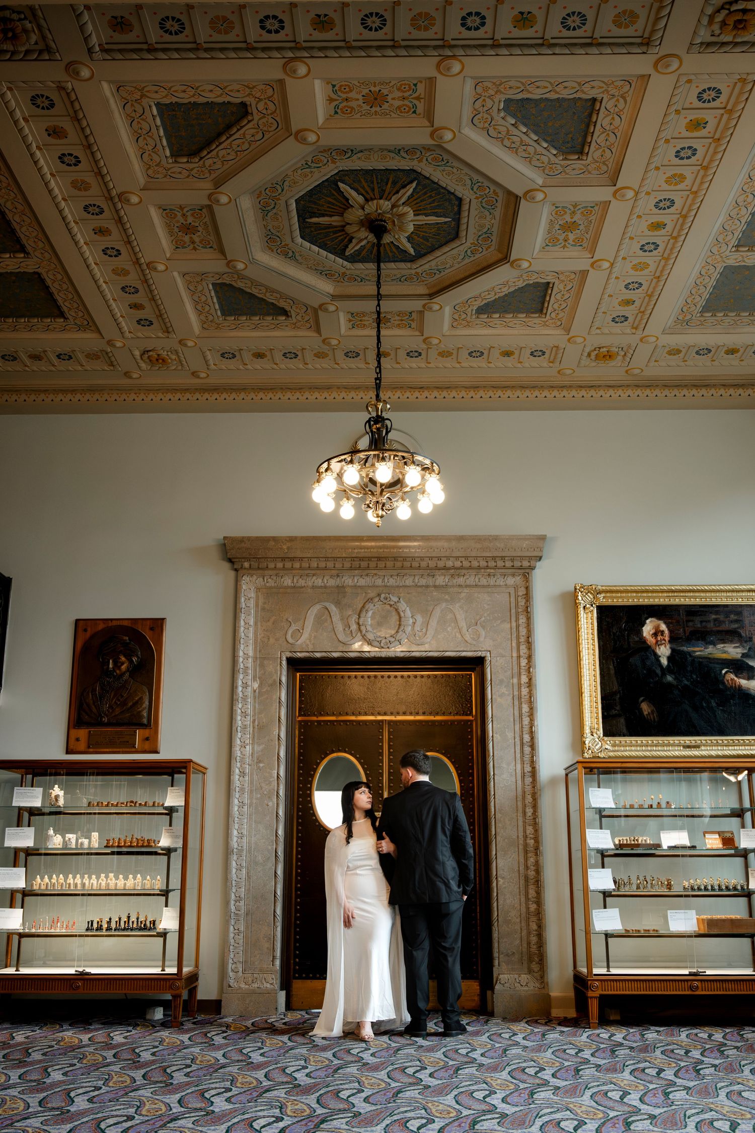 Couple in wedding attire standing beneath ornate ceiling in elegant historic venue with display cases.