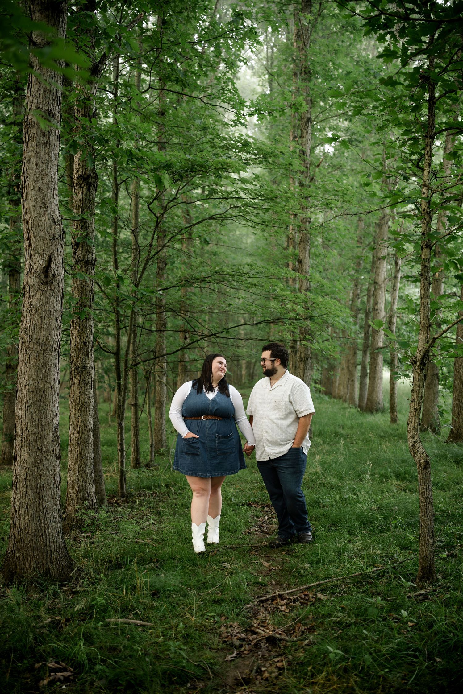 Couple holds hands walking through a lush green forest path surrounded by tall trees.