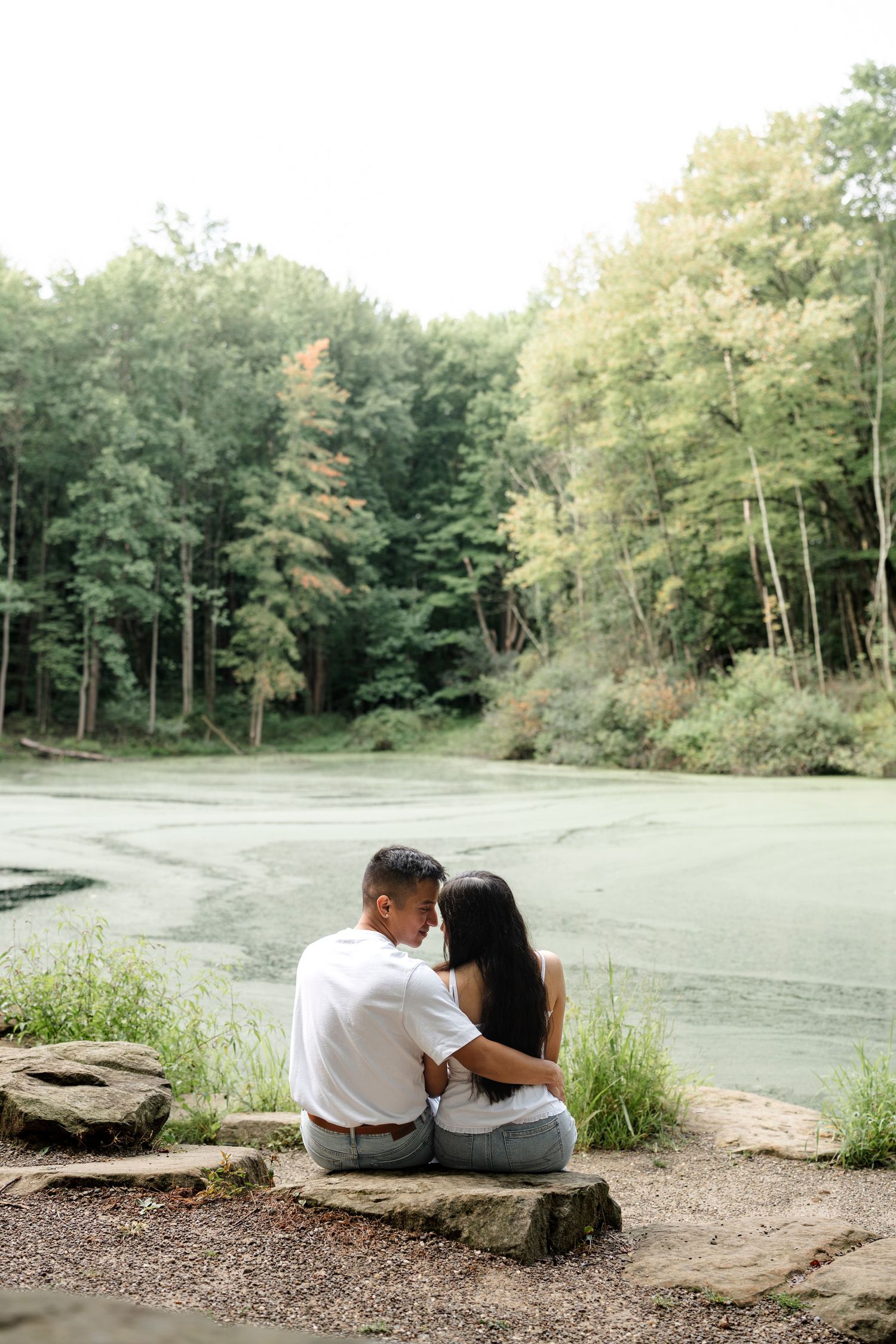 Couple sitting together on rocks by a serene green pond surrounded by lush forest trees.