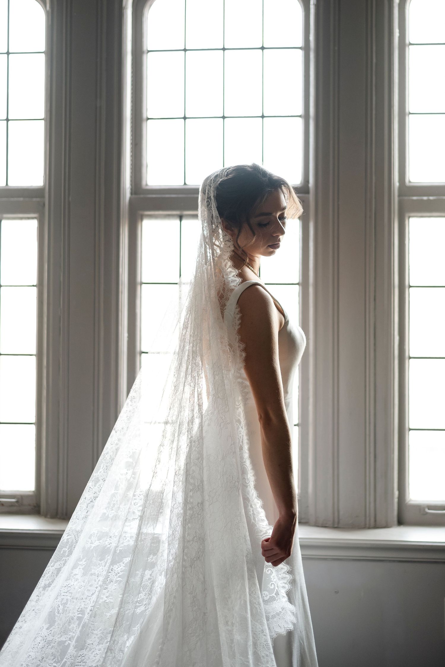 A woman in a white lace wedding dress stands in front of a large window, her veil flowing behind her.