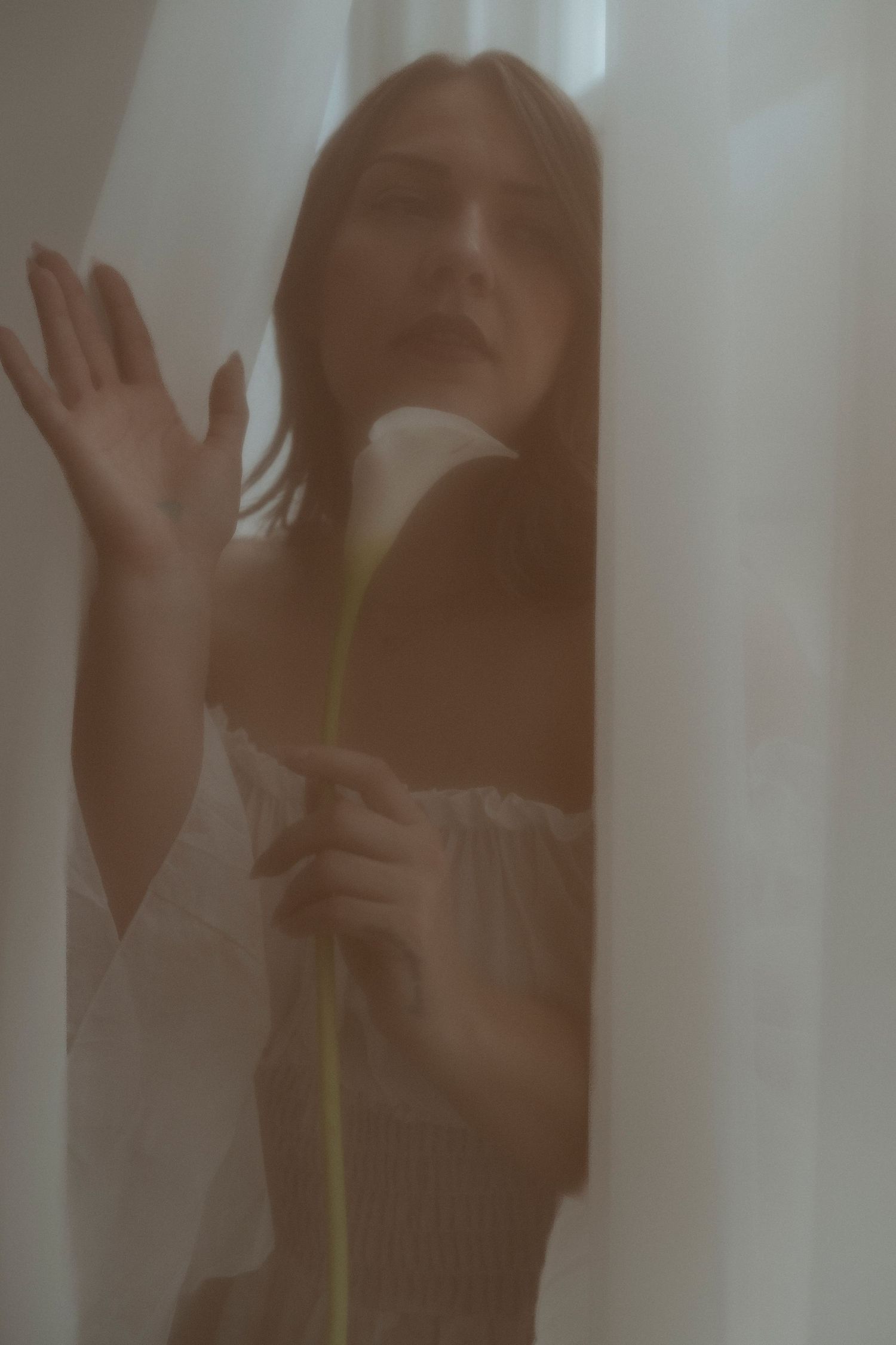 Young woman in white off-shoulder dress holding a calla lily behind sheer curtains in ethereal soft light.