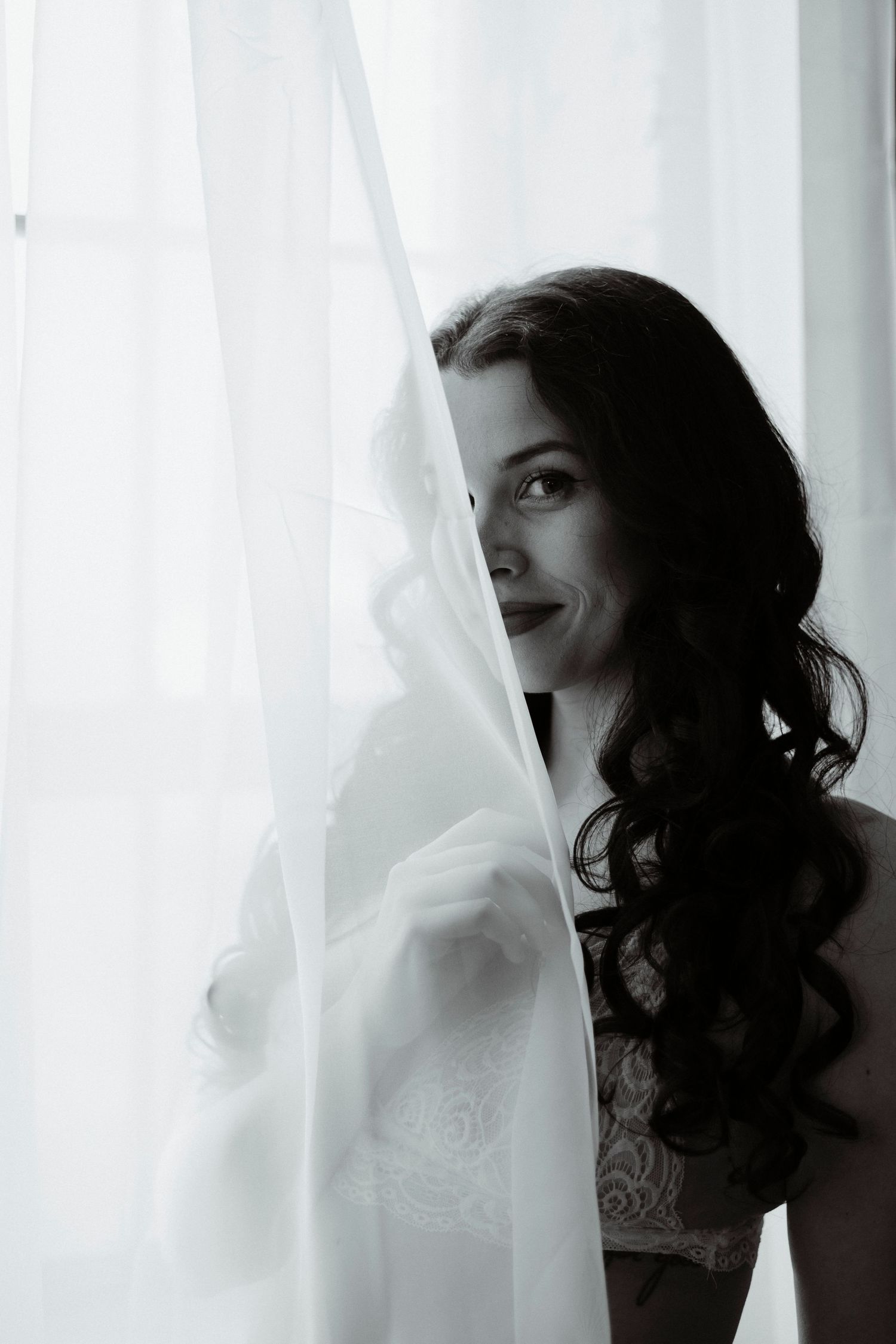 A black and white portrait of a person with dark curly hair looking through sheer white curtains in dramatic lighting.