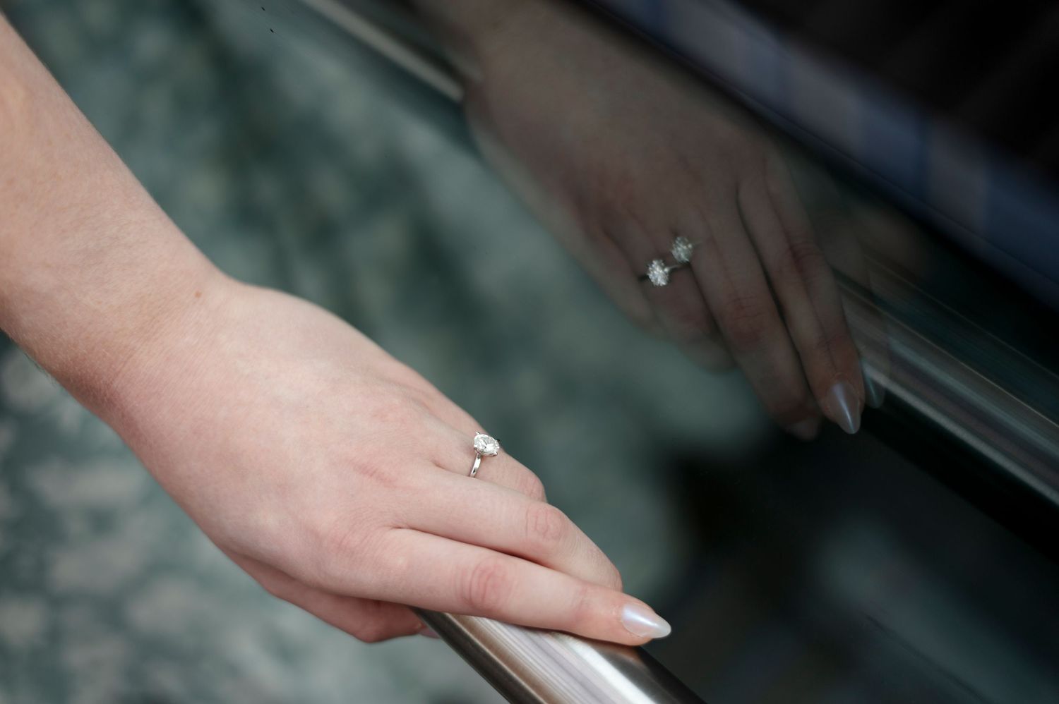 Close-up of a woman's hand wearing a diamond engagement ring, resting on a shiny chrome railing with reflection.