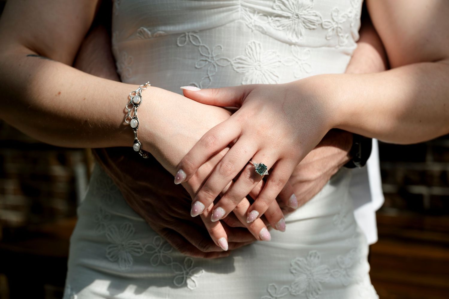 Close-up of bride and groom hands clasped together, showcasing an elegant green gemstone ring and silver bracelet.