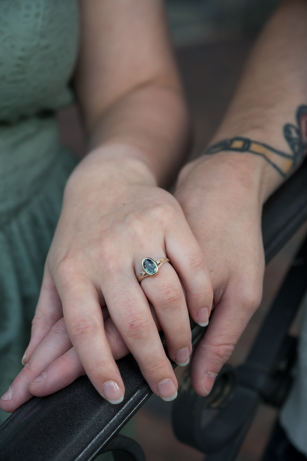 Close-up of hands showing a gold engagement ring with a blue-green gemstone, one arm featuring a tattoo.