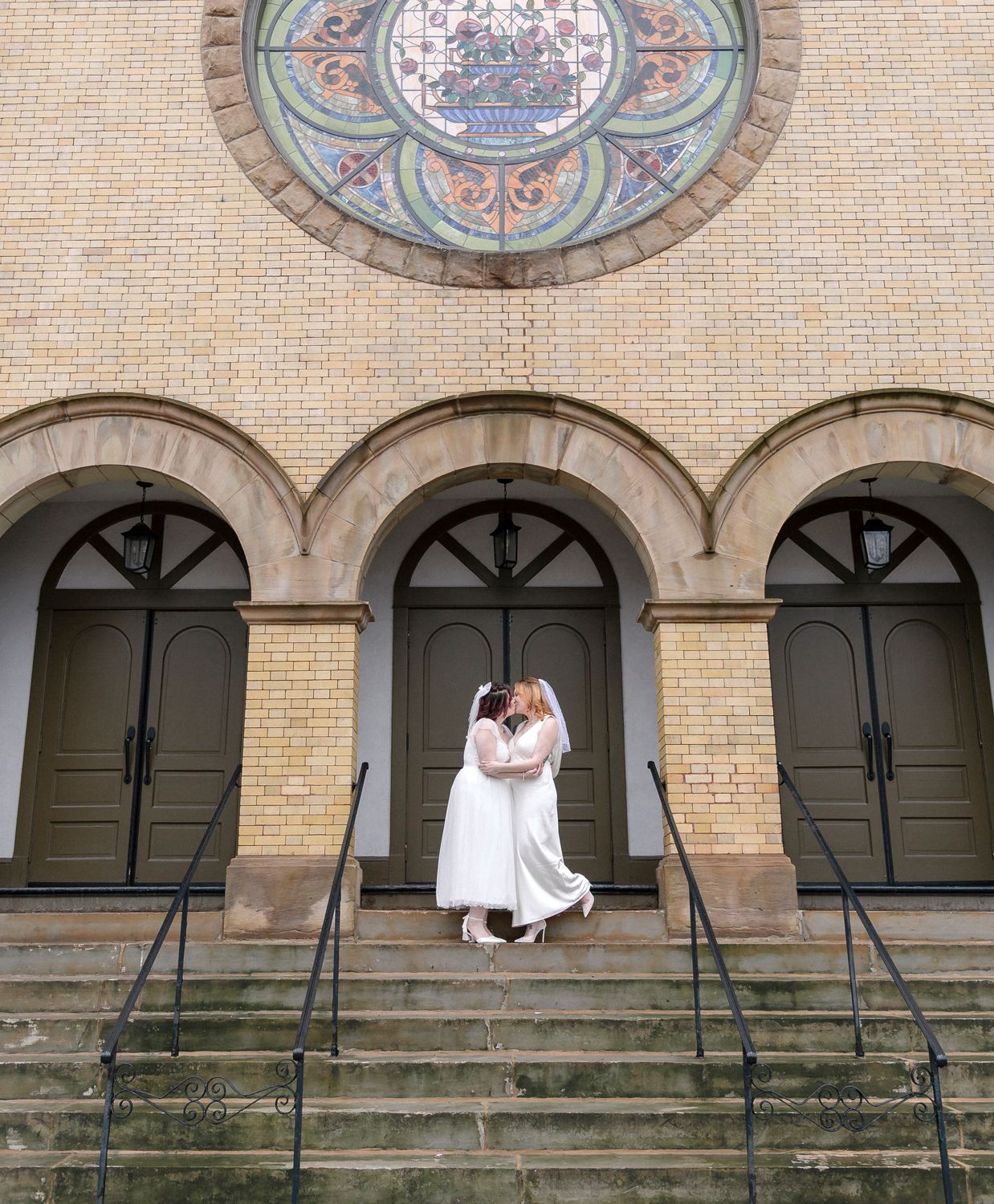 A couple in white attire photographed against an ornate brick building with arches.