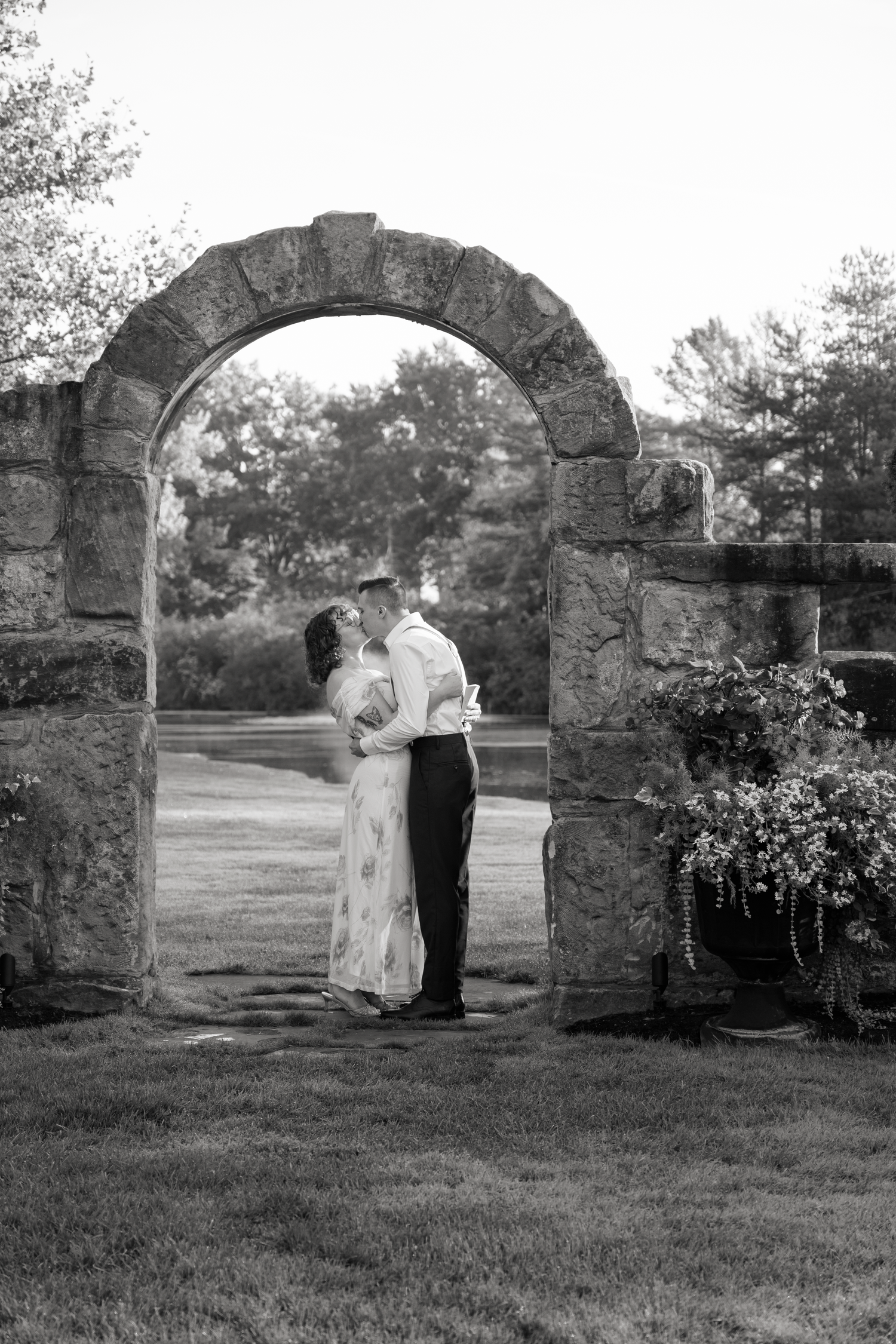 A romantic black and white photograph of a couple embracing under a rustic stone archway in a garden setting.
