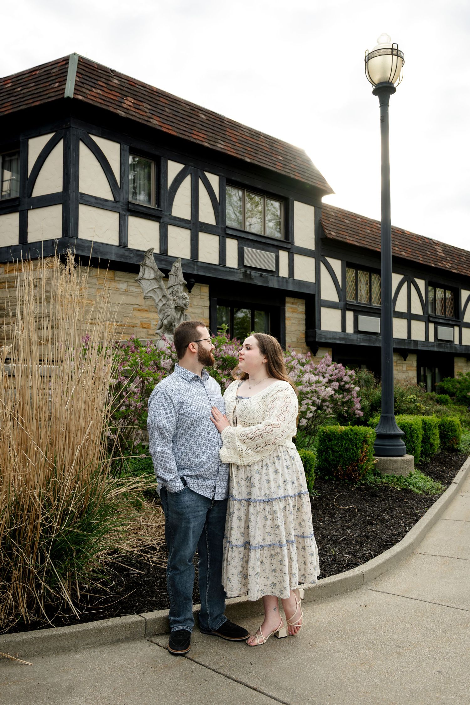 Couple standing together outside a Tudor-style building with blooming flowers and a vintage lamp post.