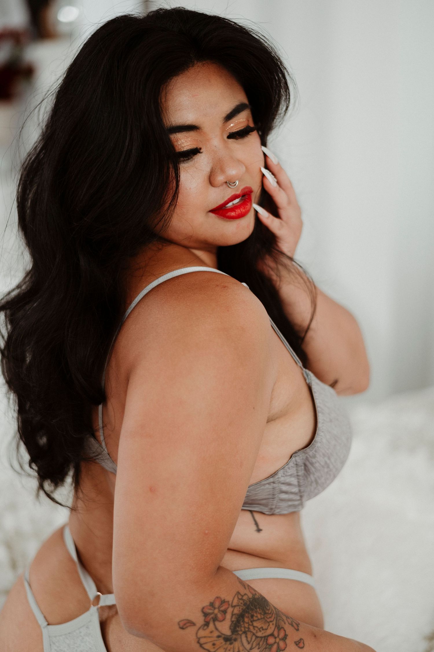 Woman with red lips and dark hair posing in gray lingerie, showcasing tattoo and bold makeup.