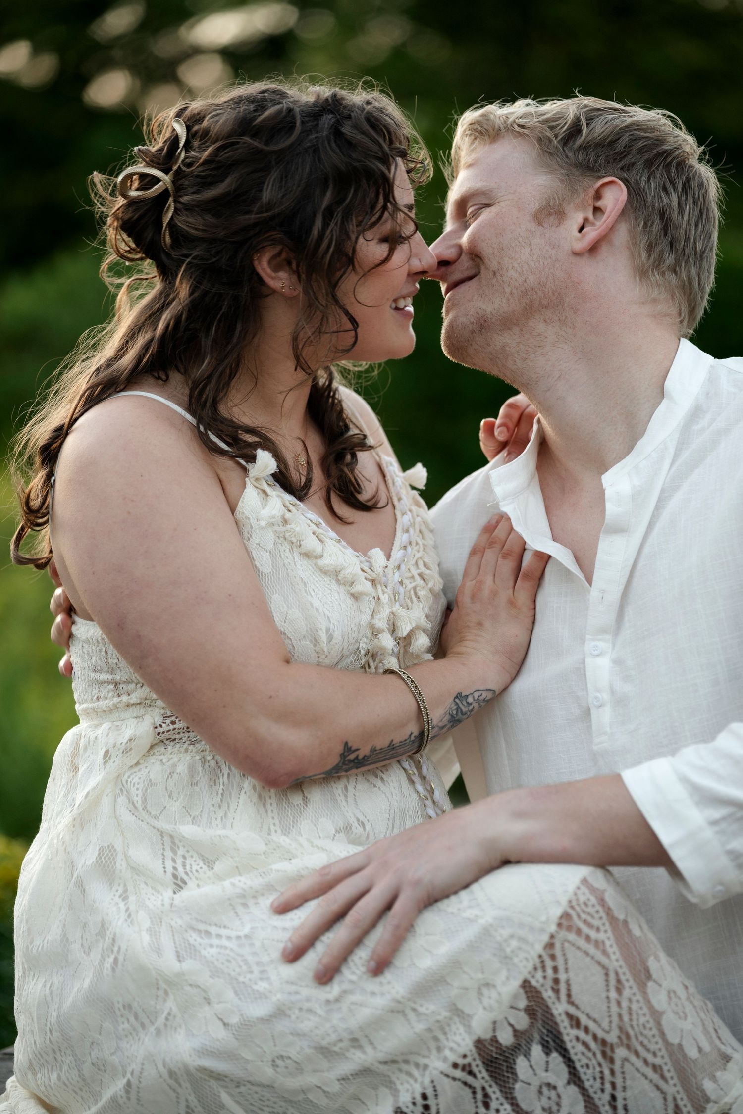 A couple in white wedding attire share a romantic moment, smiling and touching noses outdoors.