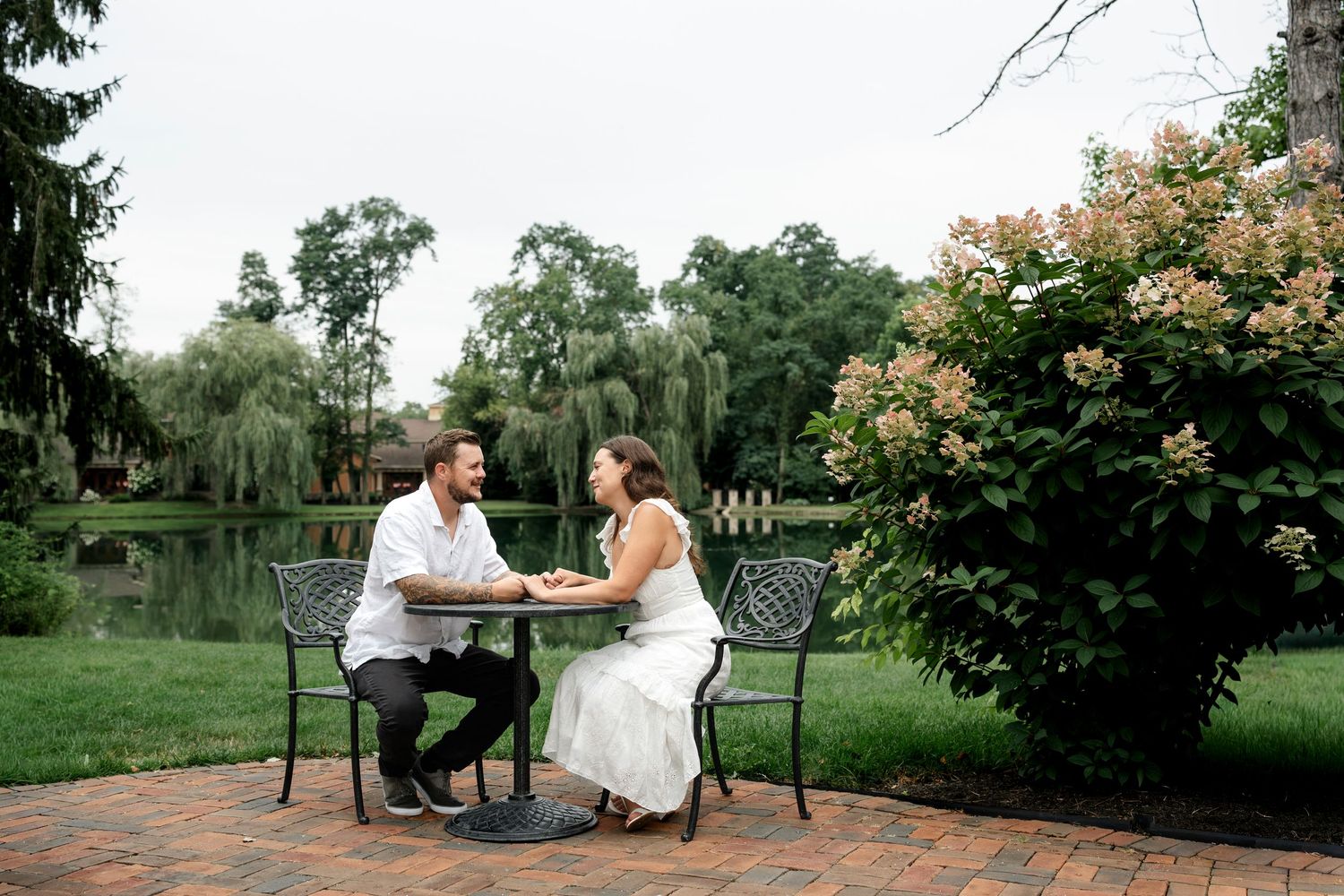 Couple in wedding attire sitting at iron table on brick patio by pond surrounded by lush greenery.
