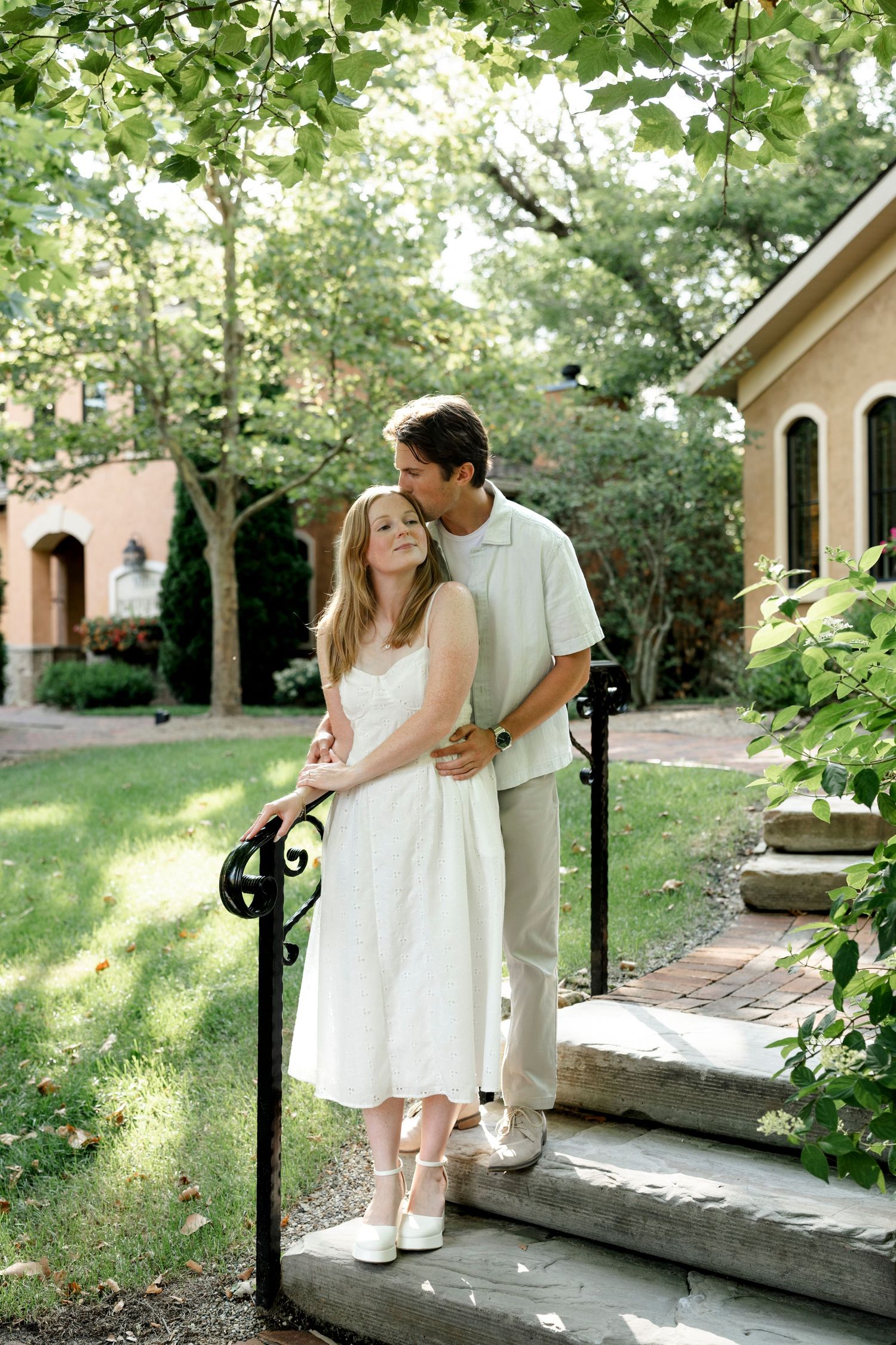 Couple embracing on garden steps, woman in white dress, man in khaki, surrounded by lush green trees near brick building.