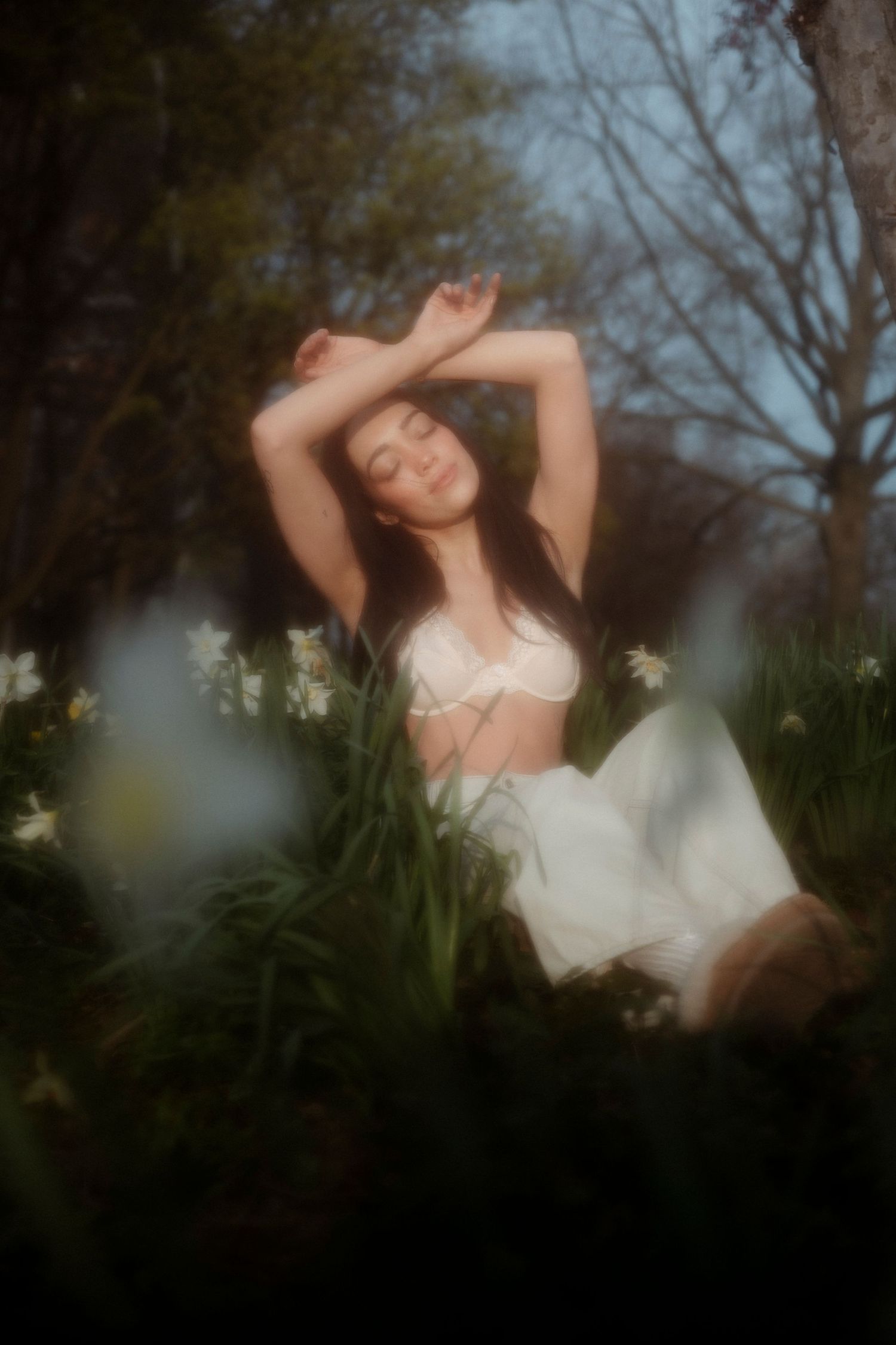 Young woman in white crop top and skirt sitting among spring flowers in a dreamy garden setting.