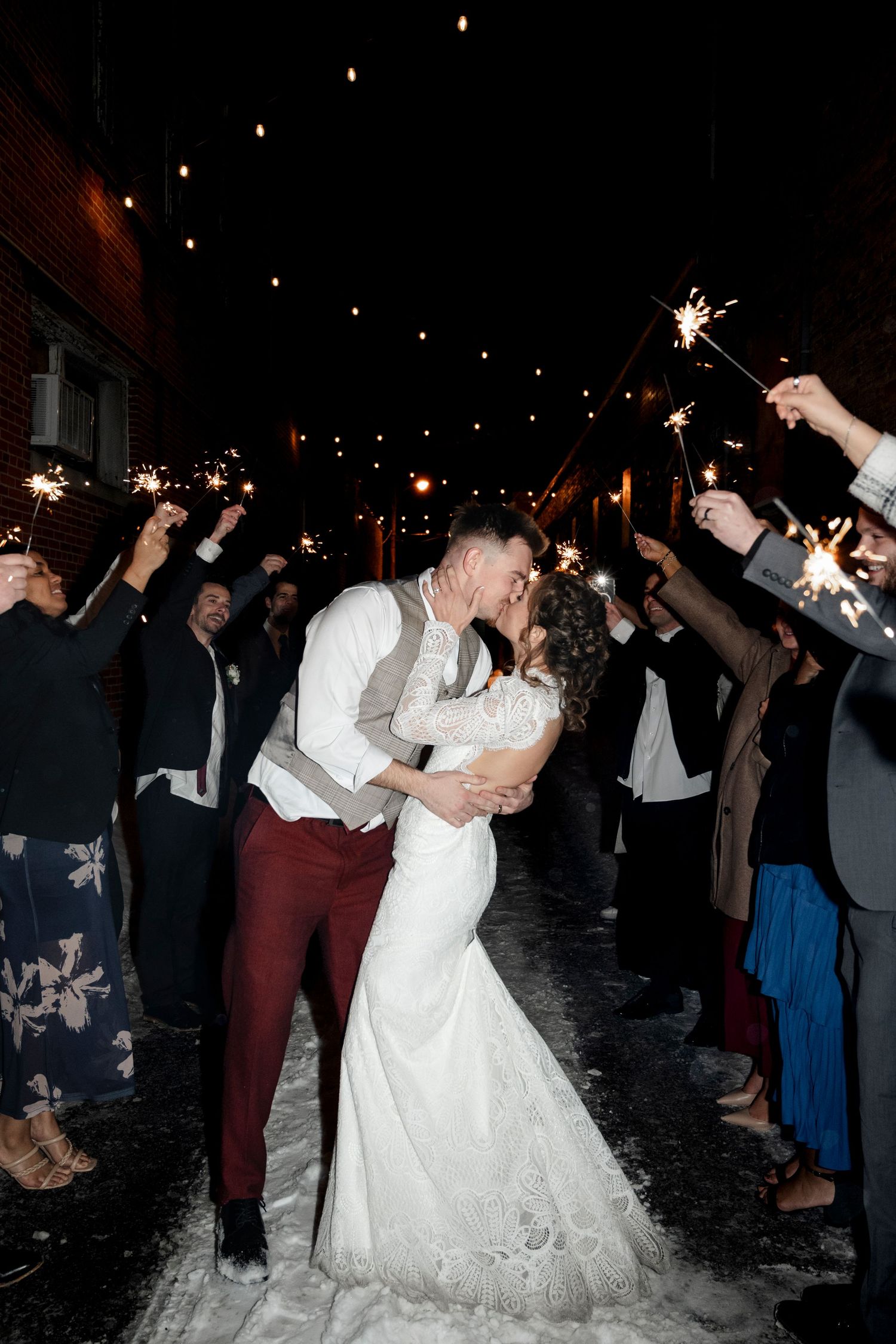 A couple embracing and kissing while surrounded by people holding sparklers in a snowy outdoor setting.