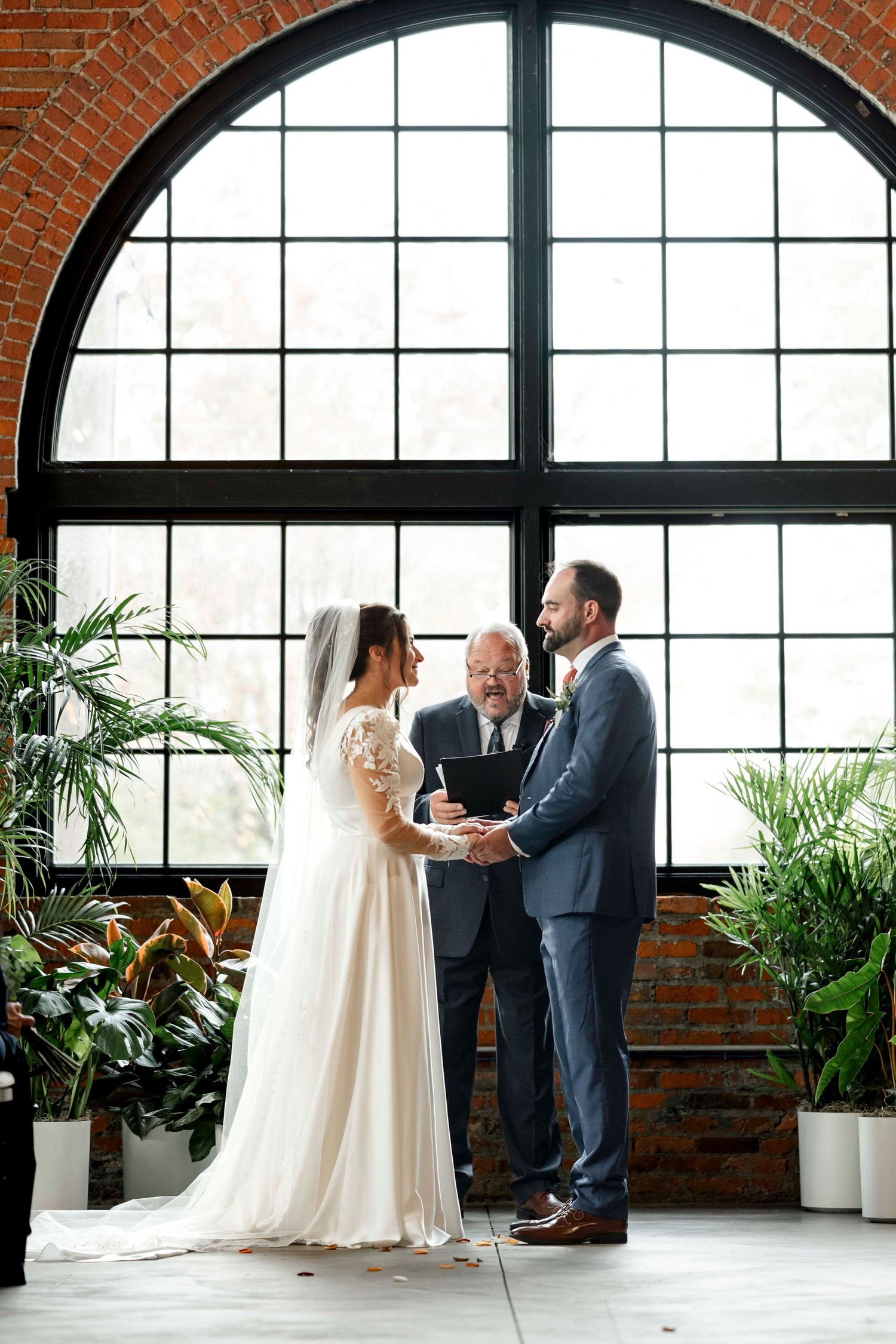 Wedding ceremony in industrial loft space with large arched window, exposed brick walls and potted plants.