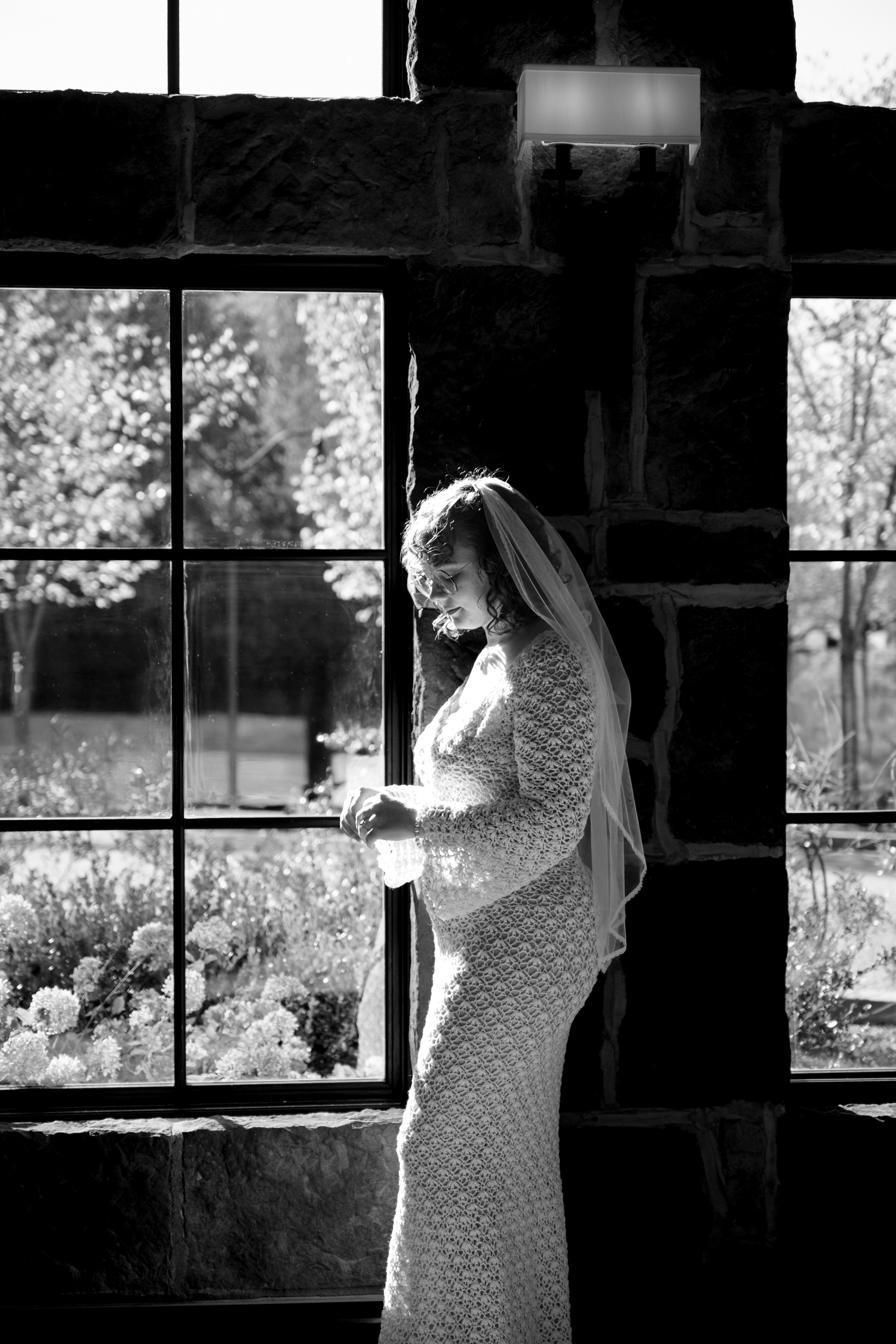 Black and white photo of bride in lace dress standing by window in dramatic lighting.
