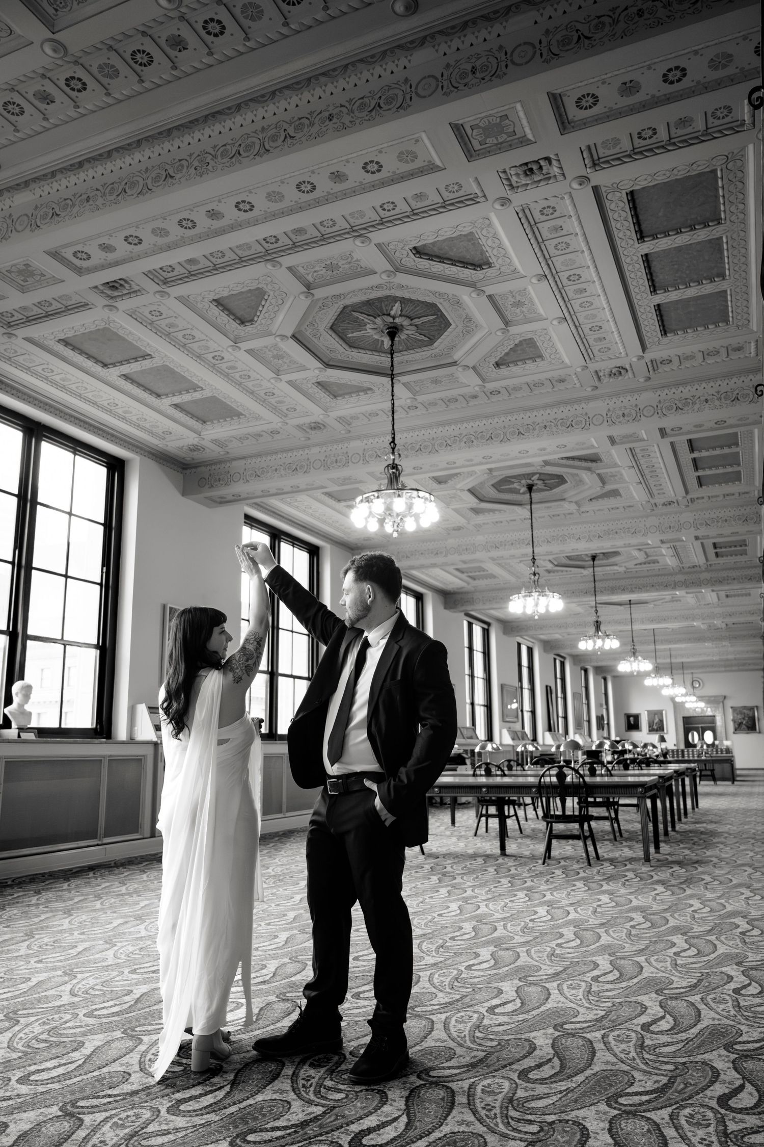 Couple dances in elegant ballroom with ornate ceiling and chandeliers in black and white photo.