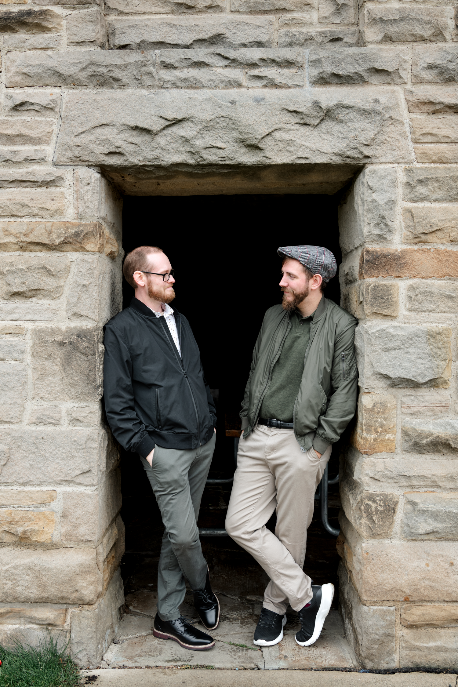 Two bearded men in casual jackets smile at each other while standing in a rustic stone doorway outdoors.