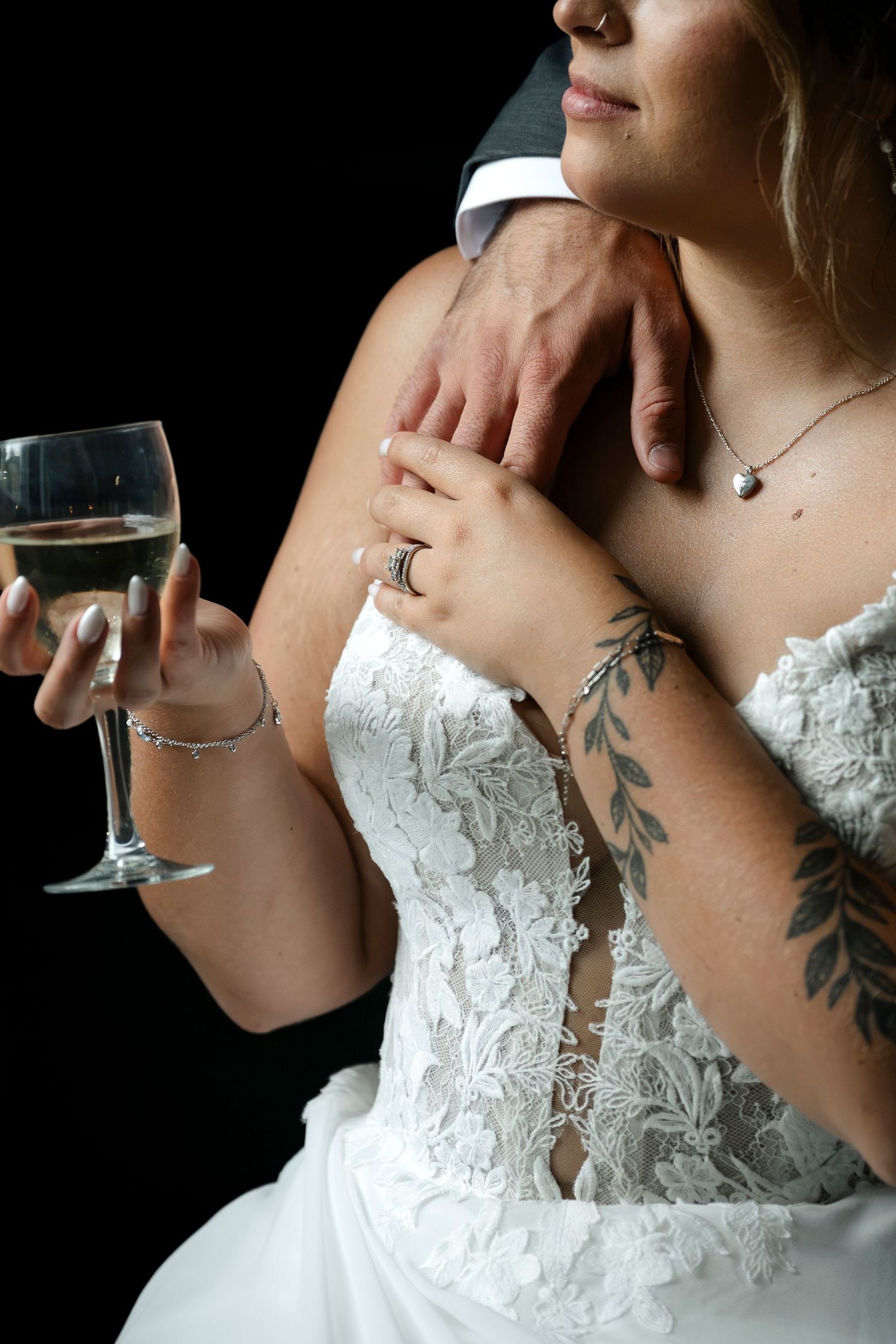Bride in lace wedding dress holding champagne glass with groom's hand on her shoulder, tattoo visible.