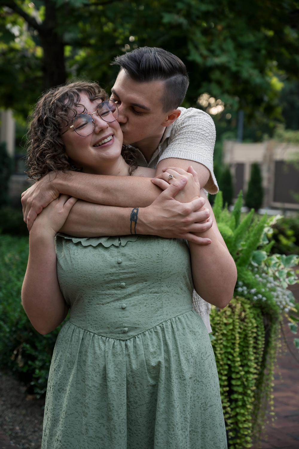 A couple embraces outdoors, man kissing woman's cheek as she smiles, surrounded by lush green garden foliage.