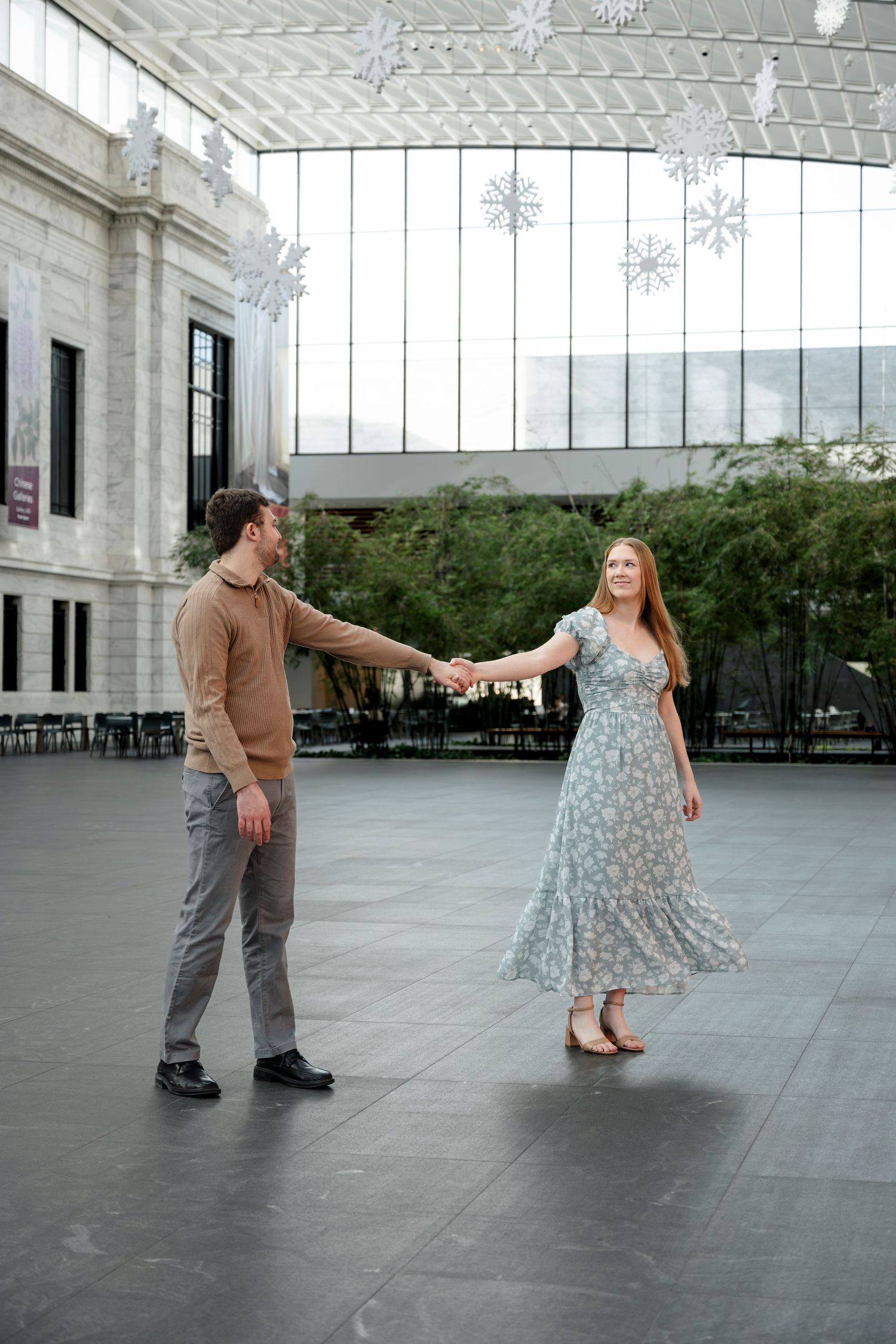 A couple holds hands and dances in an elegant indoor atrium decorated with snowflake ornaments hanging from the ceiling.