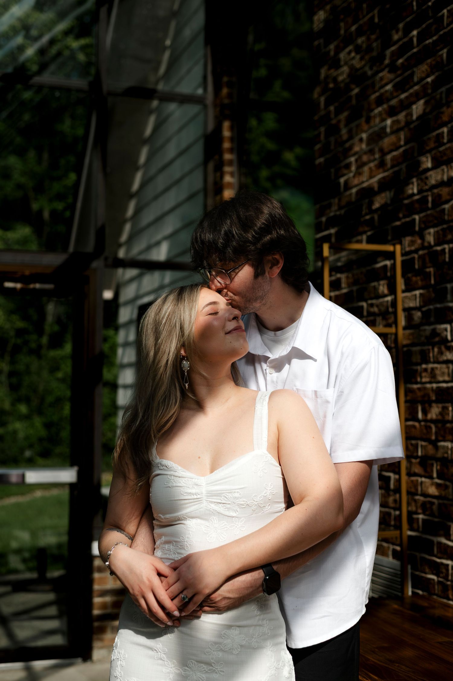 A couple in white attire shares a tender embrace near a brick wall, with the man kissing the woman's forehead.