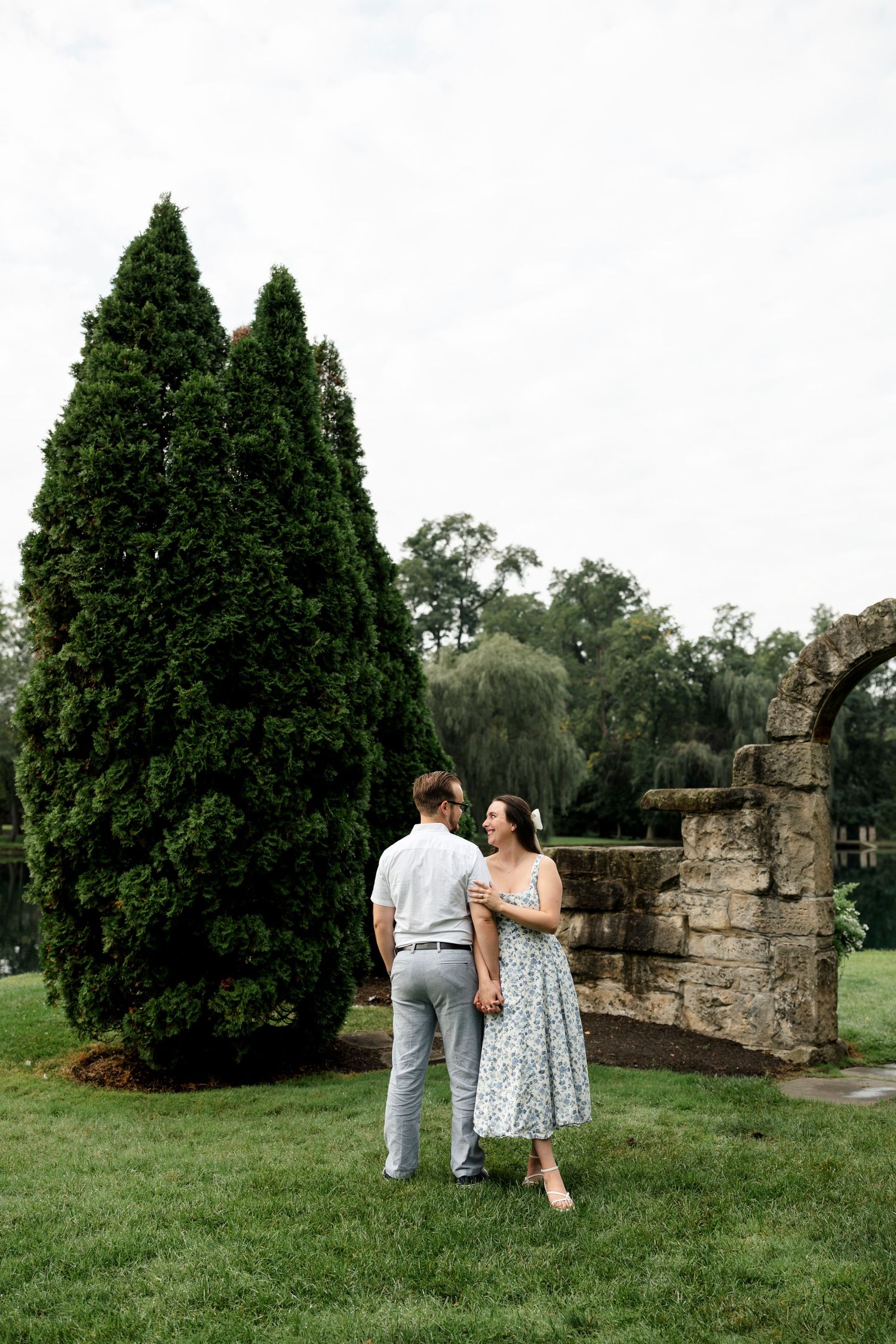 Couple holding hands near tall evergreen tree and stone ruins in a lush garden, woman in floral dress smiling.