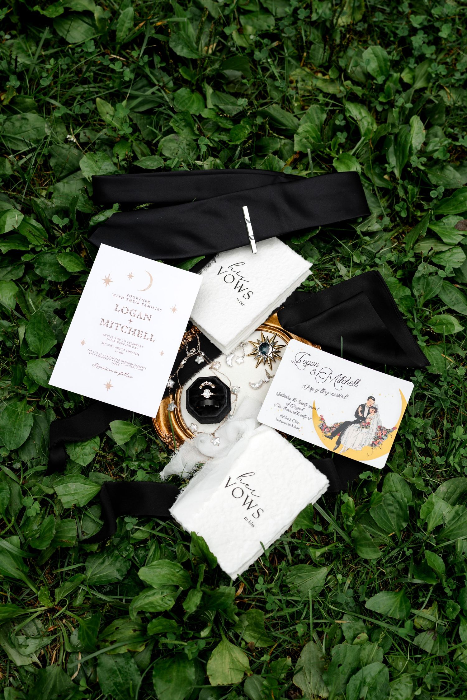 Wedding flat lay on grass featuring vow books, invitation suite, ring box, and black sash for Logan and Mitchell.