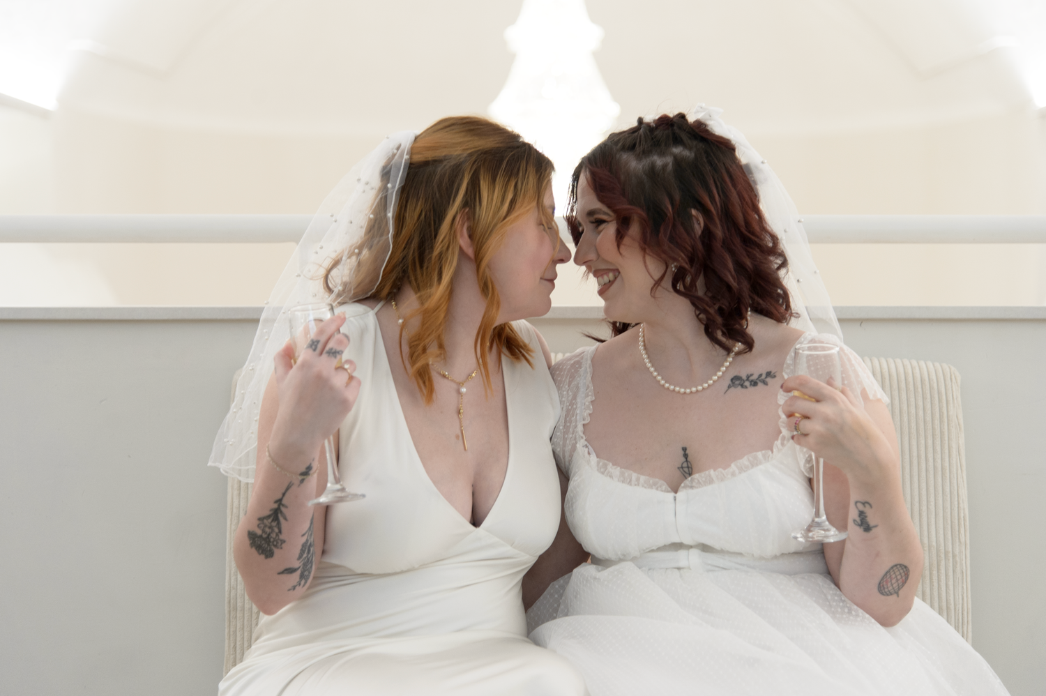 Two people in white wedding dresses lean in close together while holding champagne glasses during a celebration.