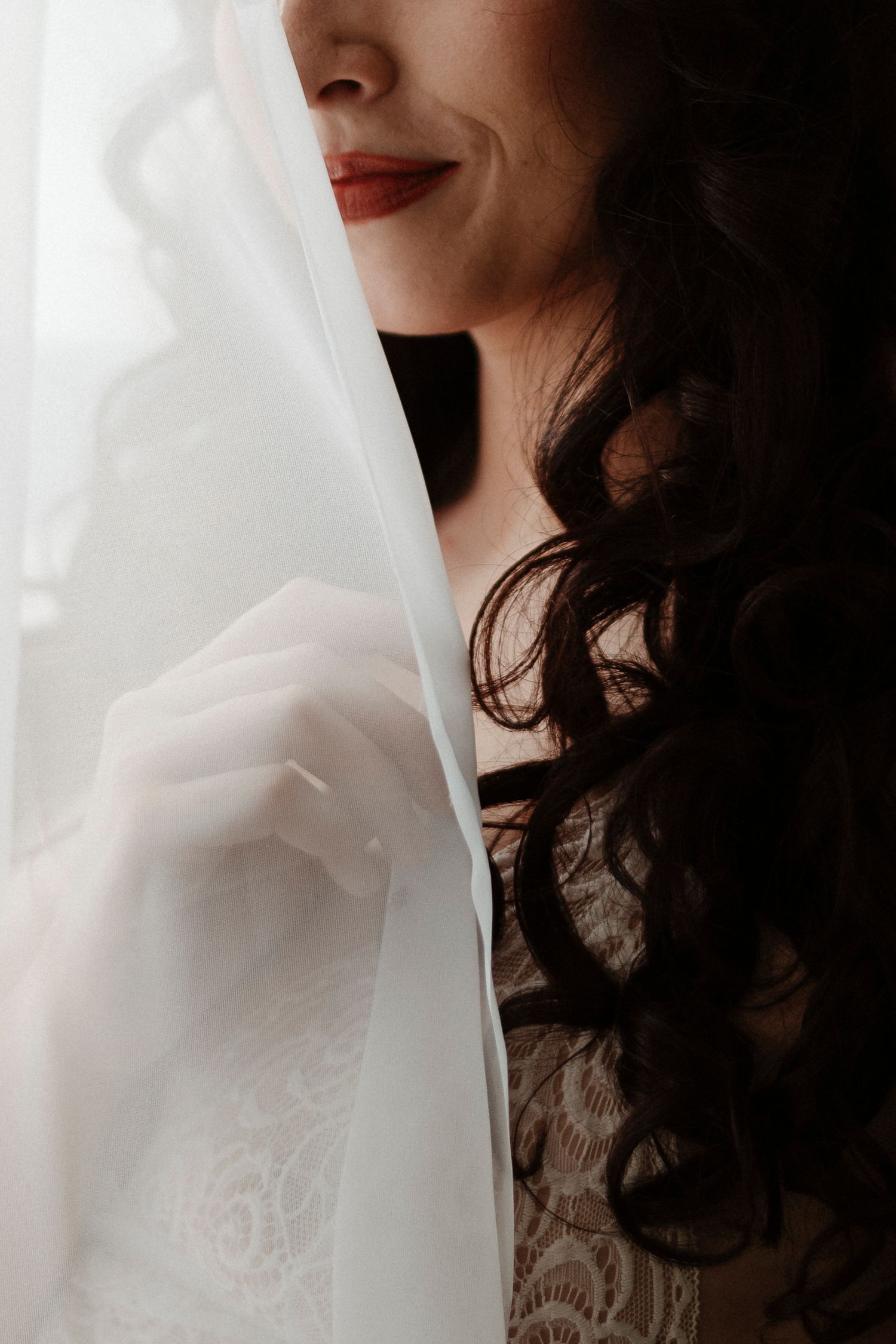A soft ethereal portrait showing dark curls and red lipstick against white tulle fabric in dramatic lighting.