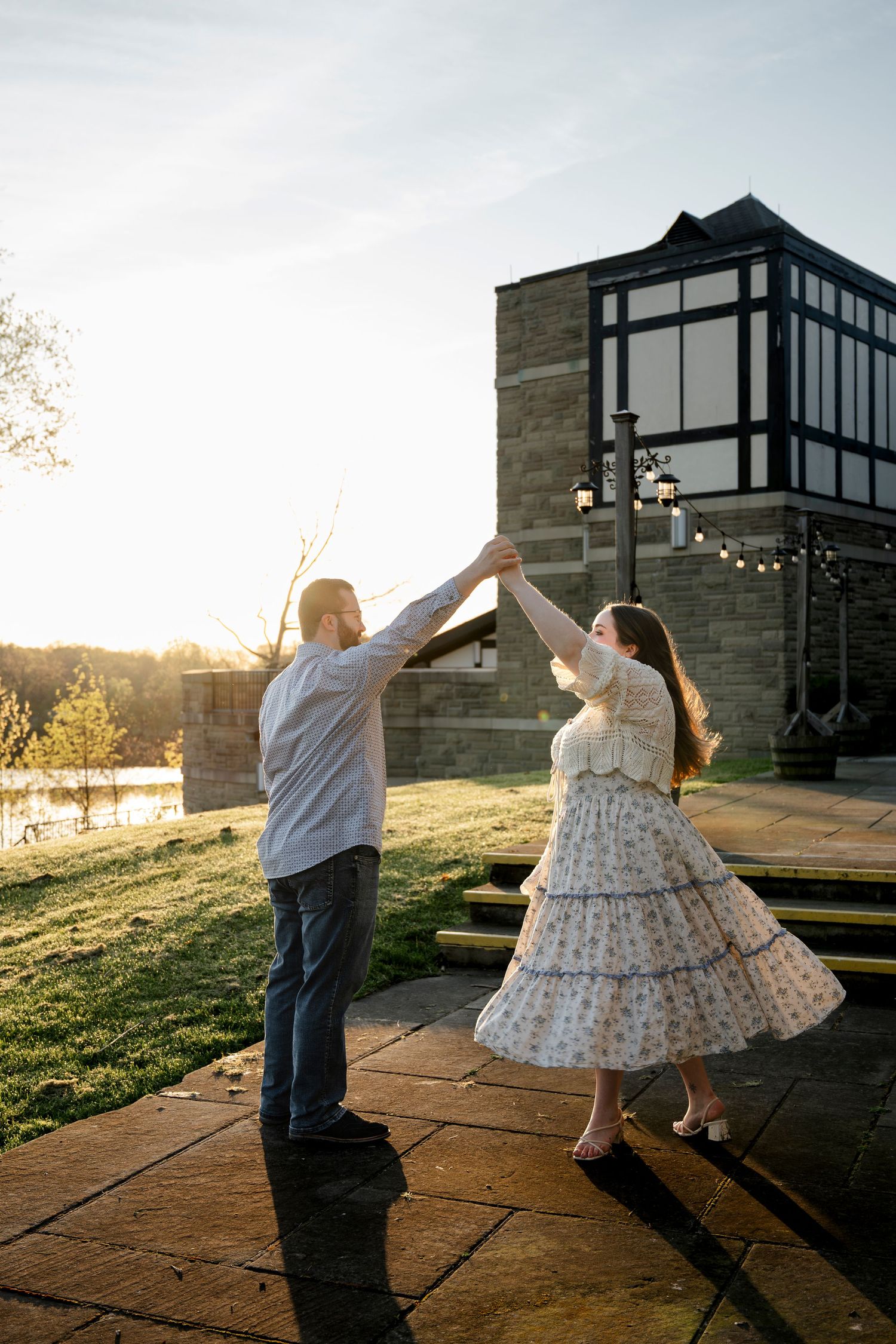 Couple dancing outdoors at golden hour near a stone building with string lights and a scenic lawn backdrop.