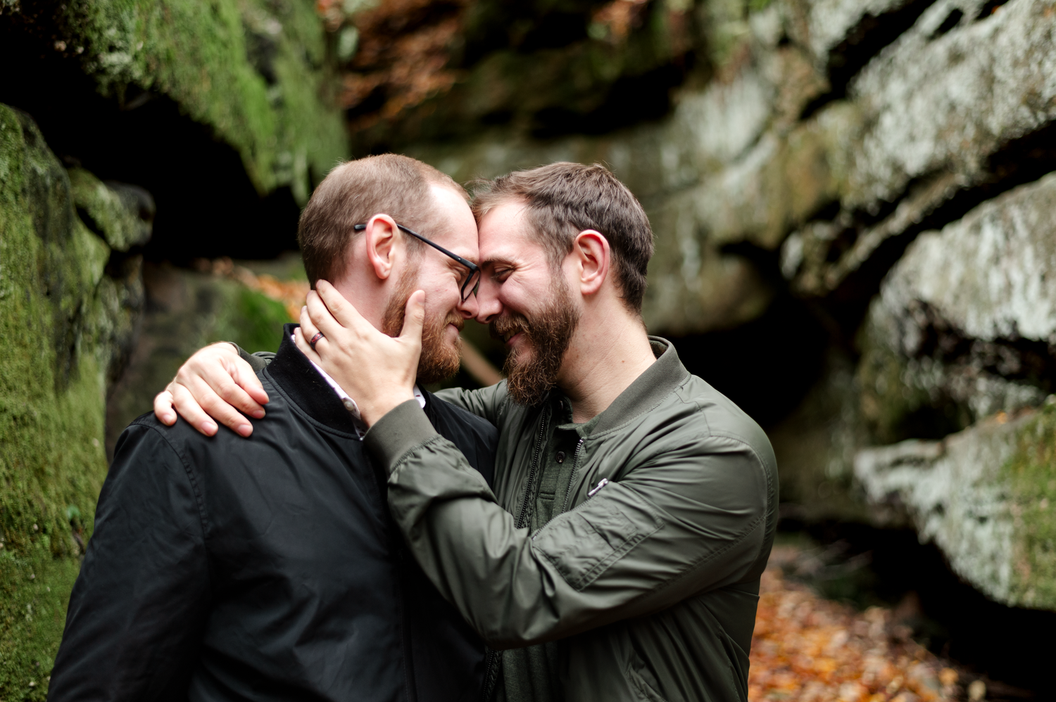 Two men sharing a tender moment, touching foreheads and embracing outdoors near rocky cliffs in autumn.