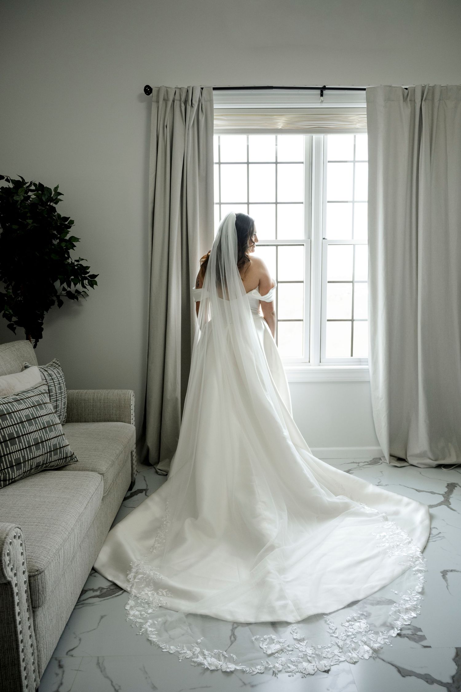 Bride in a stunning white gown with lace-trimmed cathedral veil stands before a bright window in an elegant room.