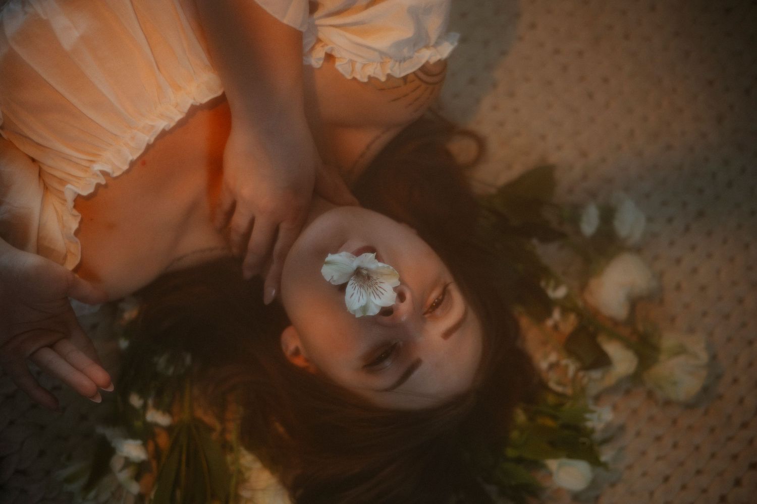Young woman lying down with white flower on face surrounded by green foliage in warm tones.