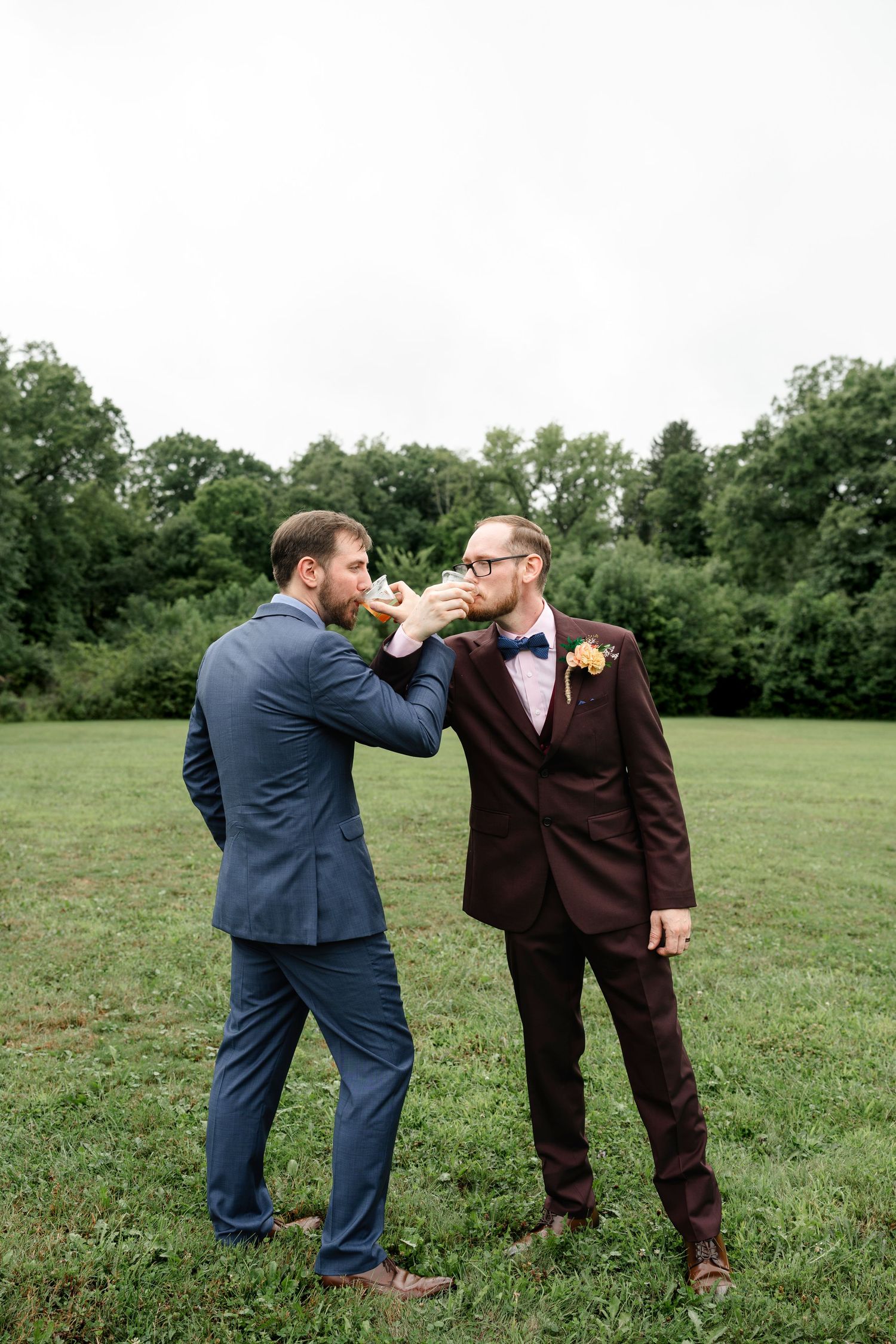 Two grooms in suits share a kiss at outdoor wedding ceremony on green grass field.