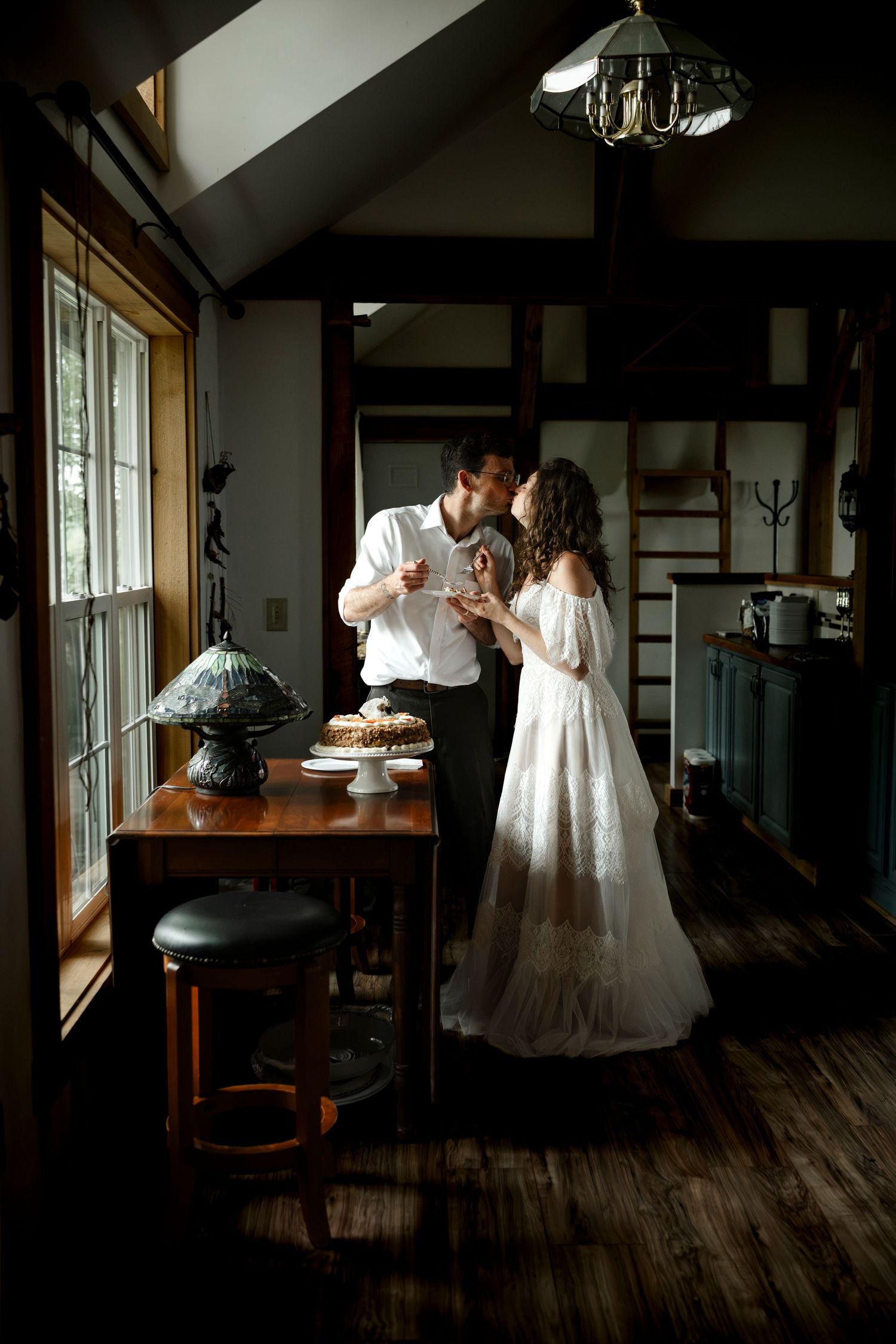 Bride and groom share a kiss while cutting their wedding cake in a rustic cabin dining room.